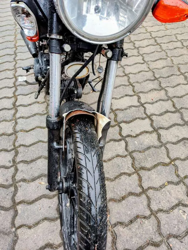 Zbliżenie przedniej części motocykla z dużym reflektorem, mniejszym światłem po lewej stronie, czarnym kołem z widocznym profilem opony i częściowo zardzewiałym błotnikiem. Motocykl stoi na brukowanej powierzchni z kamiennymi płytami ułożonymi w geometryczny wzór. Widoczne są metalowe widełki przednie oraz elementy mechanizmu.