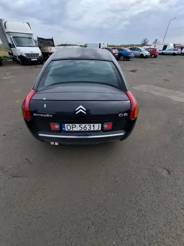 Z tyłu czarny samochód Citroën C6 z tablicą rejestracyjną OP 5631J. W tle parking z innymi pojazdami, w tym ciężarówkami i samochodami osobowymi. Niebo jest zachmurzone, a powierzchnia parkingu to asfalt. Na bagażniku widoczne logo Citroën oraz oznaczenie C6.