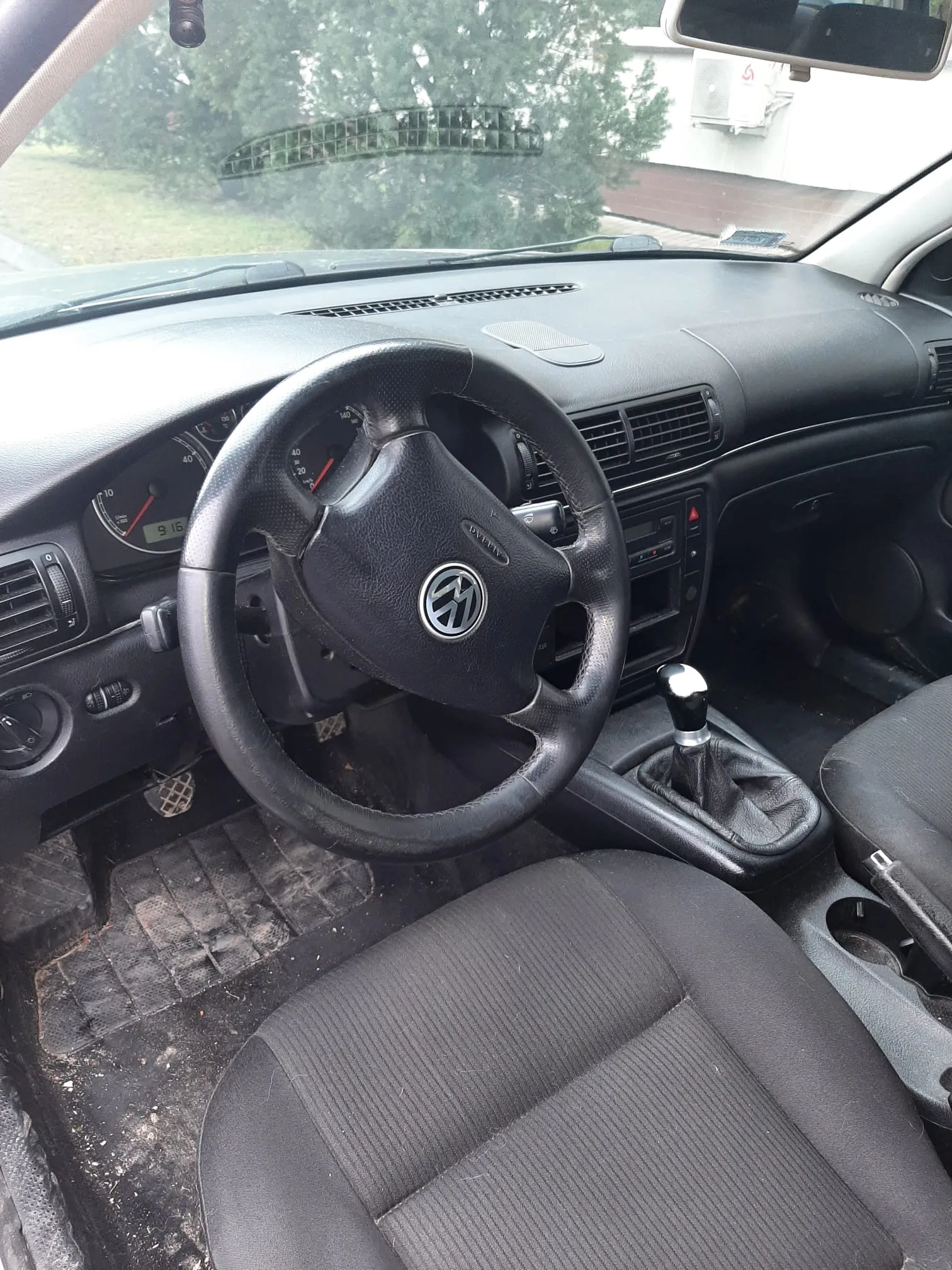 Wnętrze samochodu z czarną kierownicą z logo Volkswagen, tablicą rozdzielczą z licznikiem prędkości i cyfrowym wyświetlaczem pokazującym 916. Widoczny manualny selector biegów, czarne siedzenia tapicerowane tkaniną oraz zabrudzone dywany podłogowe. Przez przednią szybę widoczne drzewa i budynek z klimatyzatorem.