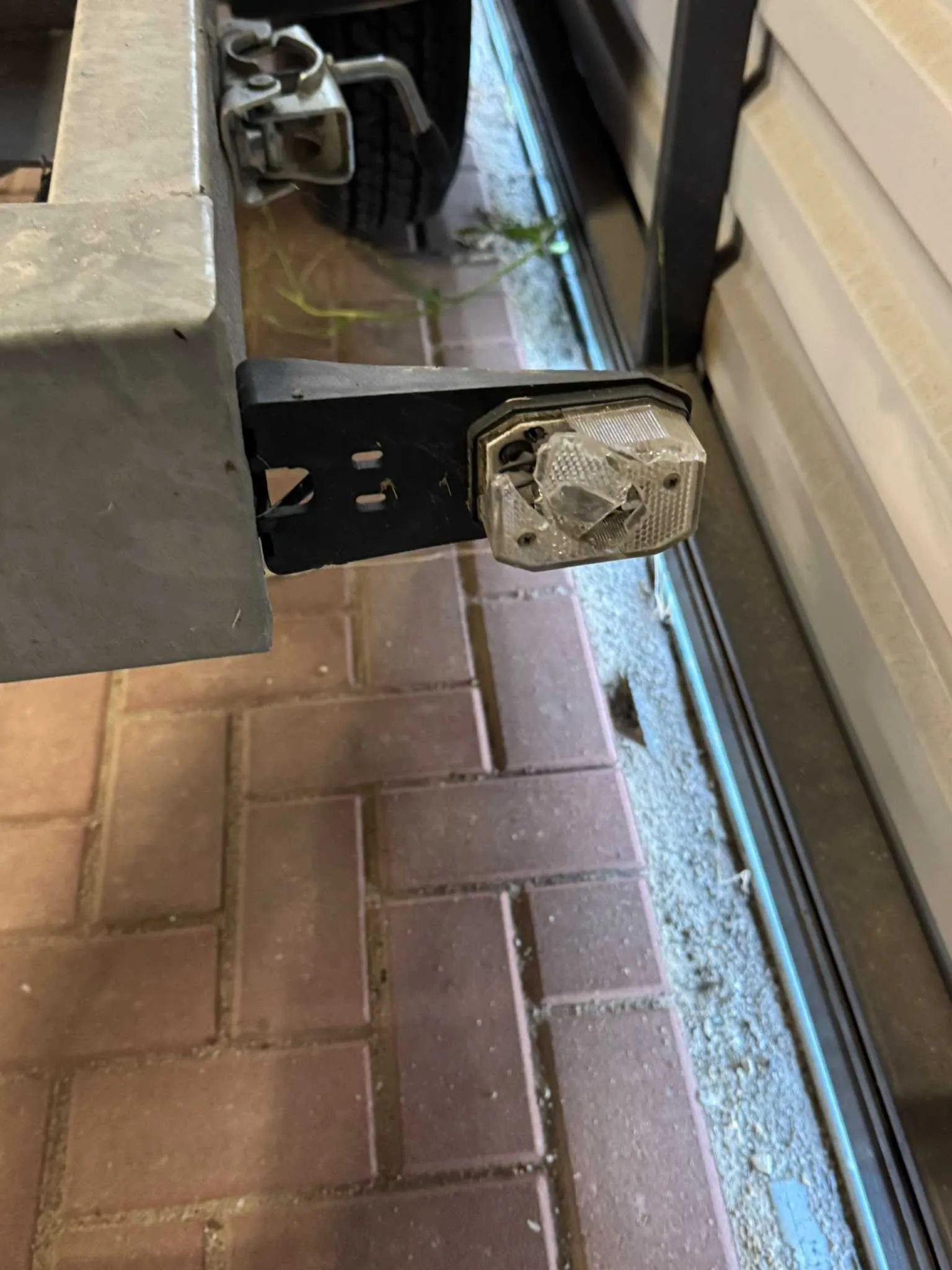 Zbliżenie na uszkodzoną tylną lampę przyczepy. Plastikowa obudowa lampy jest rozbita i popękana. Lampa jest zamocowana do czarnego, metalowego wspornika, który z kolei jest przytwierdzony do szarej, metalowej ramy przyczepy. W tle widoczna jest nawierzchnia z czerwonej kostki brukowej oraz fragment ściany budynku.