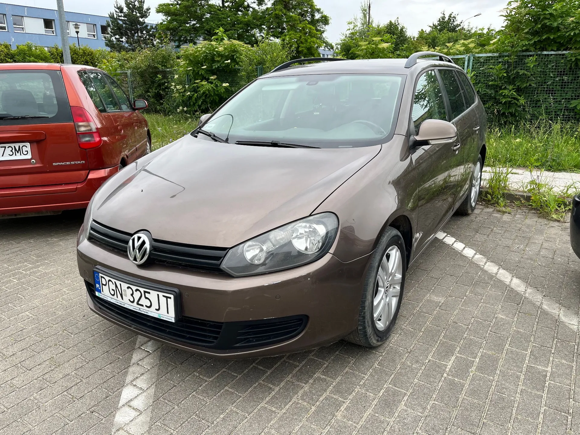 Brązowy samochód osobowy marki Volkswagen w nadwoziu kombi zaparkowany na kostce brukowej. Samochód ma polską tablicę rejestracyjną o numerze PGN 325JT. Obok, po lewej stronie, widoczny jest fragment czerwonego samochodu marki Mitsubishi Space Star z tablicą kończącą się na 73MG. W tle znajdują się zielone drzewa, krzewy oraz fragmenty budynków mieszkalnych.