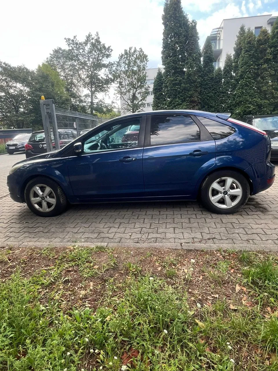 Profil granatowego samochodu osobowego typu hatchback zaparkowanego na kostce brukowej. W tle widoczne są inne samochody, zielone drzewa oraz fragment białego budynku mieszkalnego. Na pierwszym planie znajduje się pas trawy i ziemi.