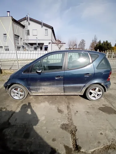Niebieski samochód osobowy typu hatchback zaparkowany na betonowej powierzchni z widocznymi plamami wilgoci. Samochód jest zabrudzony, szczególnie na dolnej części karoserii. W tle budynek mieszkalny z białym ogrodzeniem, drzewa bez liści oraz jasne niebo z nielicznymi chmurami. Na betonie widoczny cień osoby robiącej zdjęcie.