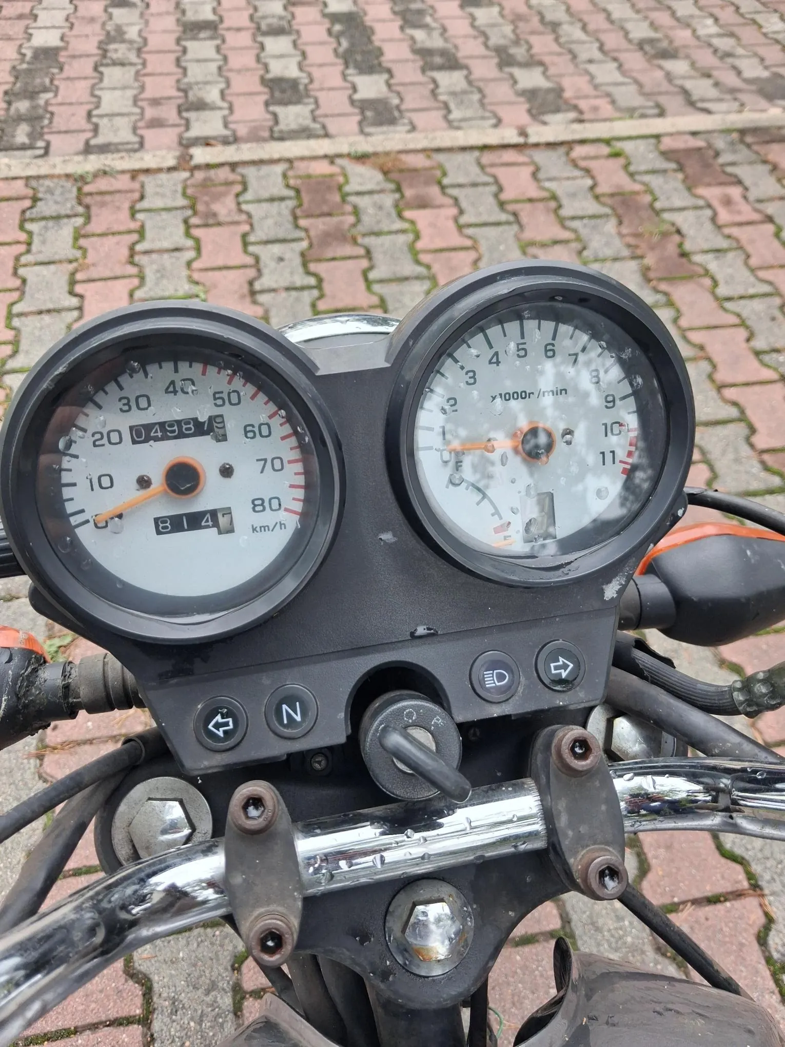 Zbliżenie na deskę rozdzielczą motocykla. Widoczne są dwa okrągłe analogowe wskaźniki: po lewej prędkościomierz z zaznaczoną prędkością do 100 km/h i licznikiem kilometrów wskazującym 04987, po prawej obrotomierz z oznaczeniem x1000r/min. Poniżej wskaźników znajduje się panel z przyciskami, w tym wskaźnik neutralnego biegu 'N' oraz ikona świateł. Całość zamontowana jest na chromowanej kierownicy, a w tle widoczna jest kostka brukowa w kolorach szarym i czerwonym.