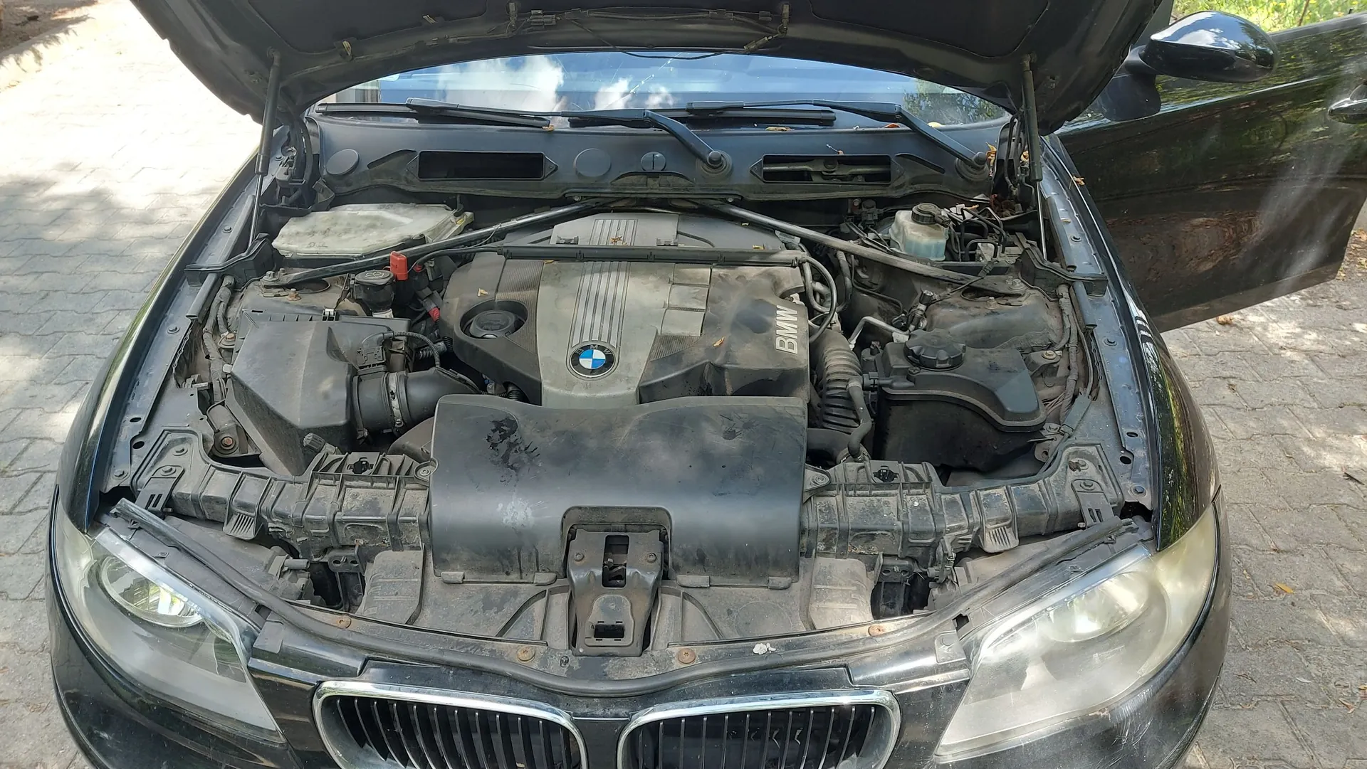 Widok z góry na otwartą maskę czarnego samochodu marki BMW, ukazującą zakurzony silnik i elementy komory silnika. Na plastikowej osłonie silnika widoczne jest logo BMW.