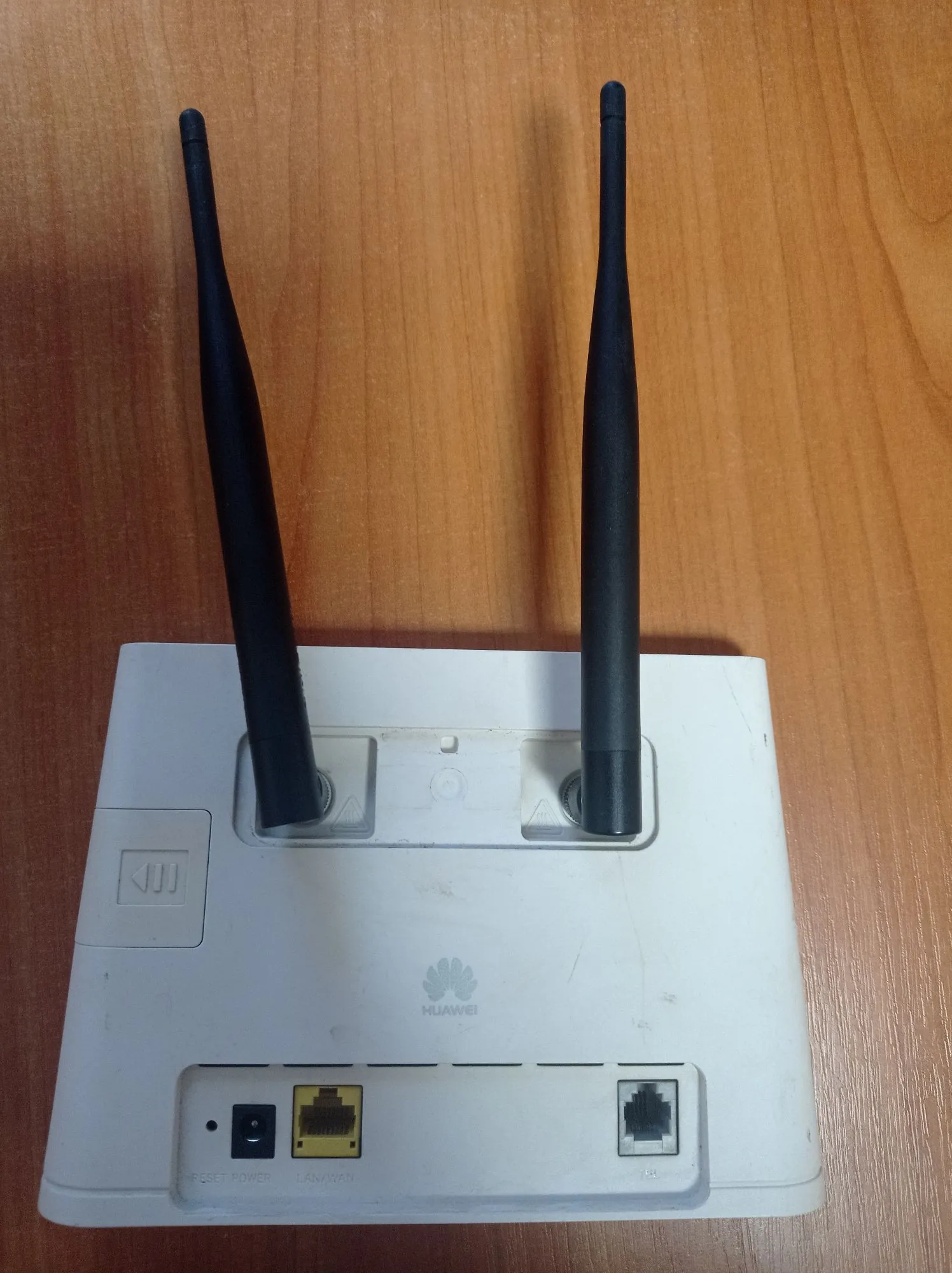Biały router marki Huawei z dwiema czarnymi antenami skierowanymi w górę, ustawiony na drewnianym blacie. Na przednim panelu urządzenia widoczne jest logo Huawei oraz gniazda: zasilania, port LAN/WAN oraz port telefoniczny RJ11. Po lewej stronie znajduje się mała naklejka z ikoną.