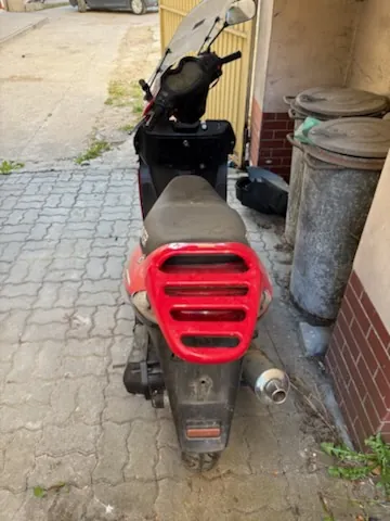 Czerwono-czarny skuter zaparkowany tyłem do obserwatora na brukowanej nawierzchni. Widoczne są czerwone tylne światła z poziomymi szczelinami, brudne siedzenie, wydech oraz tabliczka rejestracyjna bez czytelnego tekstu. Po prawej stronie obrazu znajdują się dwa duże, szare pojemniki na śmieci przy ceglanej ścianie budynku. W tle widoczna jest ulica z fragmentem innego pojazdu i chodnikiem.