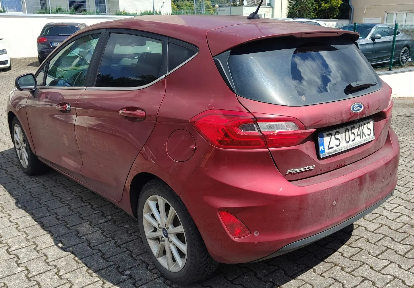Czerwony samochód marki Ford Fiesta zaparkowany na kostce brukowej. Widok z tyłu i z boku pojazdu. Samochód posiada polską tablicę rejestracyjną o numerze ZS 054KS.