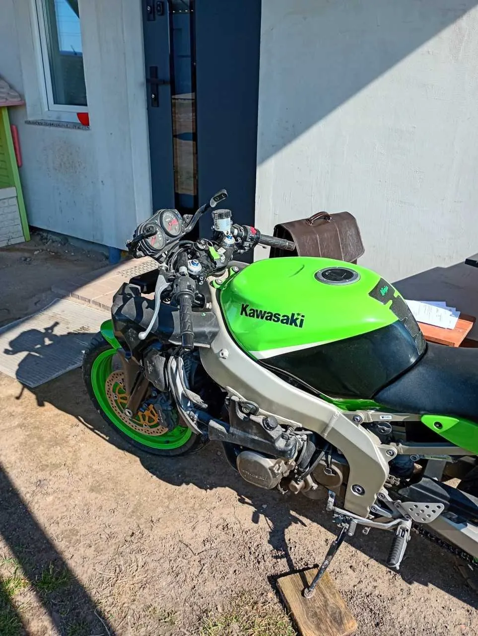 Zdjęcie motocykla marki Kawasaki o kolorze zielonym i srebrnym, zaparkowanego na podwórku. Widoczny napis 'Kawasaki' na zbiorniku paliwa, brązowa torba na siedzeniu, tablica przyrządów oraz elementy mechanizmu. W tle budynek z niebieską drzwiami i oknem, na ziemi leżą dokumenty na drewnianym stole. Motocykl oparty jest na podstawce z drewnianym blokiem.