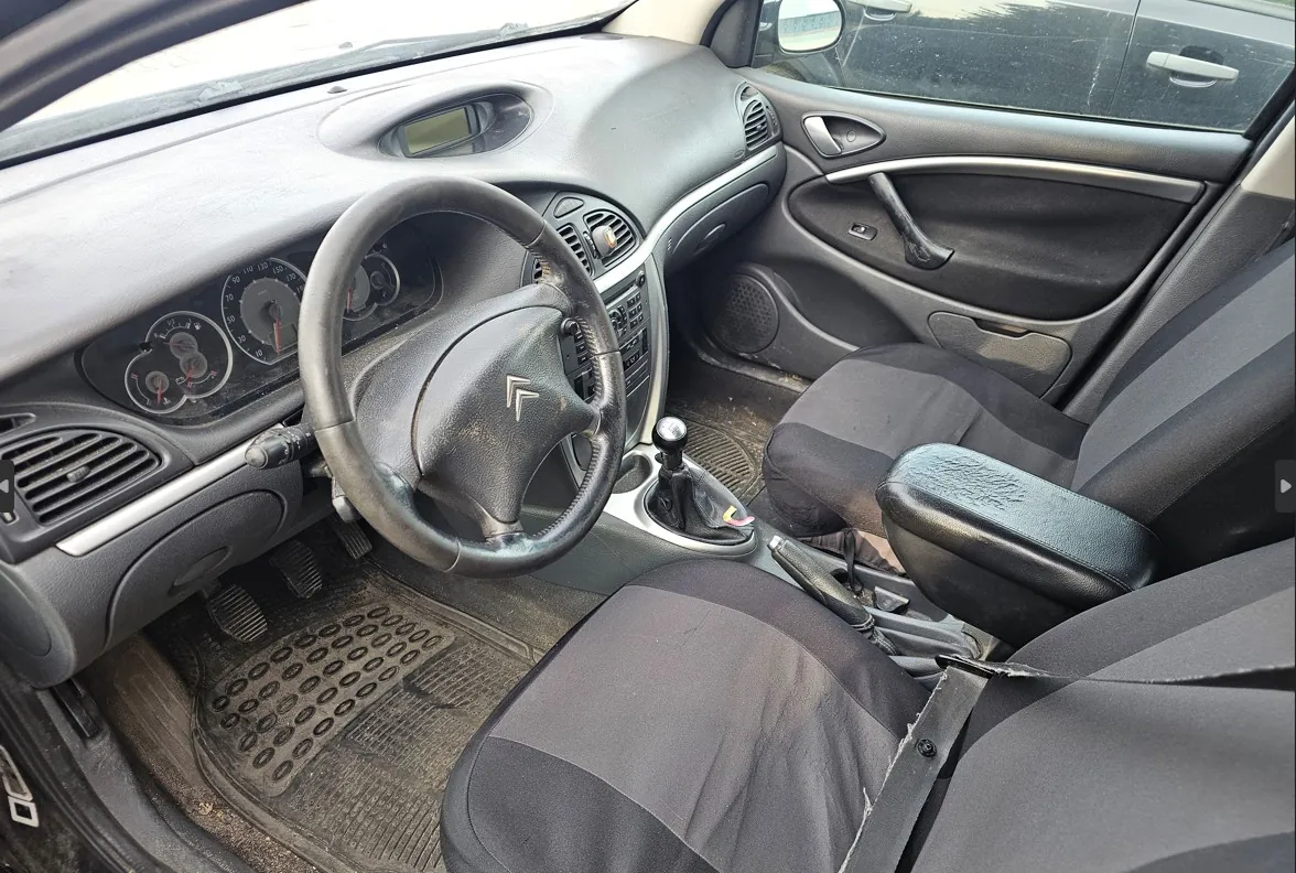 Wnętrze samochodu z perspektywy kierowcy. Widoczna czarna kierownica z logo Citroëna, deska rozdzielcza z analogowymi wskaźnikami prędkości i obrotów, lewarek zmiany biegów oraz dwa przednie fotele z czarnymi pokrowcami. W tle widoczny panel drzwi kierowcy oraz fragment przedniej szyby.