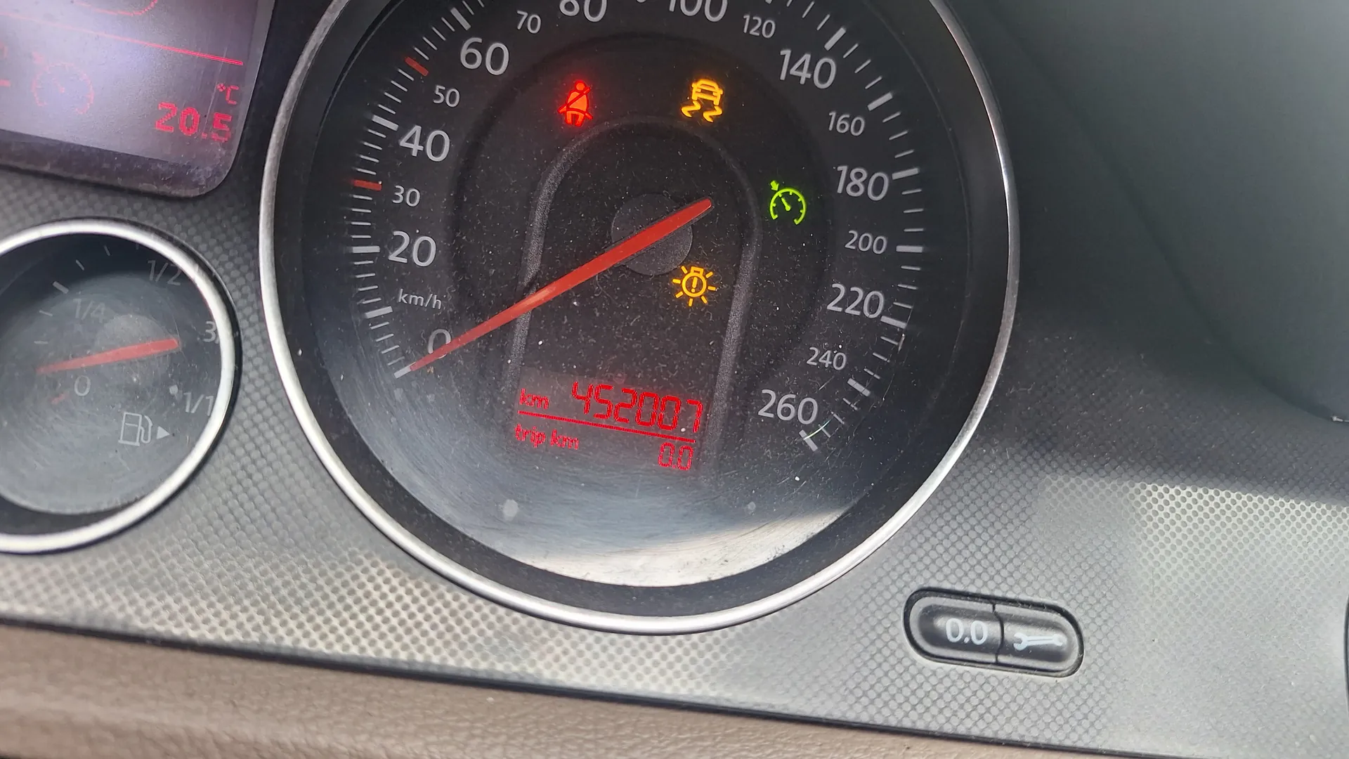 Zbliżenie na deskę rozdzielczą samochodu. W centrum znajduje się prędkościomierz z czerwoną igłą wskazującą blisko 0 km/h. Wyświetlacz cyfrowy pokazuje przebieg 45200.7 km oraz napis 'trip km'. Aktywne są żarówka ostrzegawcza (żółta), zielony symbol obrotomierza, pomarańczowy symbol silnika i czerwony symbol pasa bezpieczeństwa. Po prawej stronie widoczny wskaźnik paliwa i wyświetlacz temperatury z wartością 20.5°C.