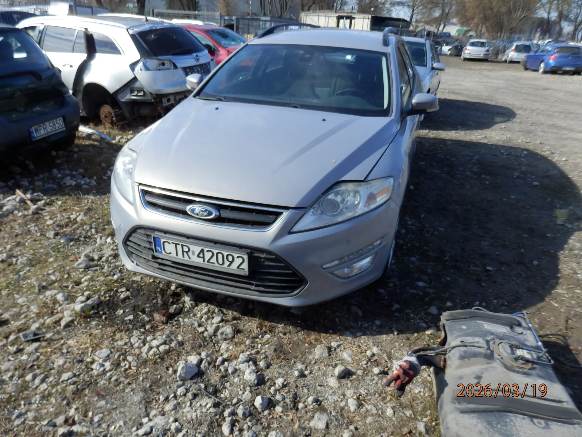 Srebrny samochód marki Ford z tablicą rejestracyjną CTR 42092 (PL) na pierwszym planie. W tle uszkodzone pojazdy, w tym biały samochód z otwartą klapą i czarny samochód z tablicą WPR 5850. Teren pokryty żwirem i kamieniami. W prawym dolnym rogu widoczna data 2026/03/19 oraz częściowo zdemontowany element pojazdu z rękawicą.