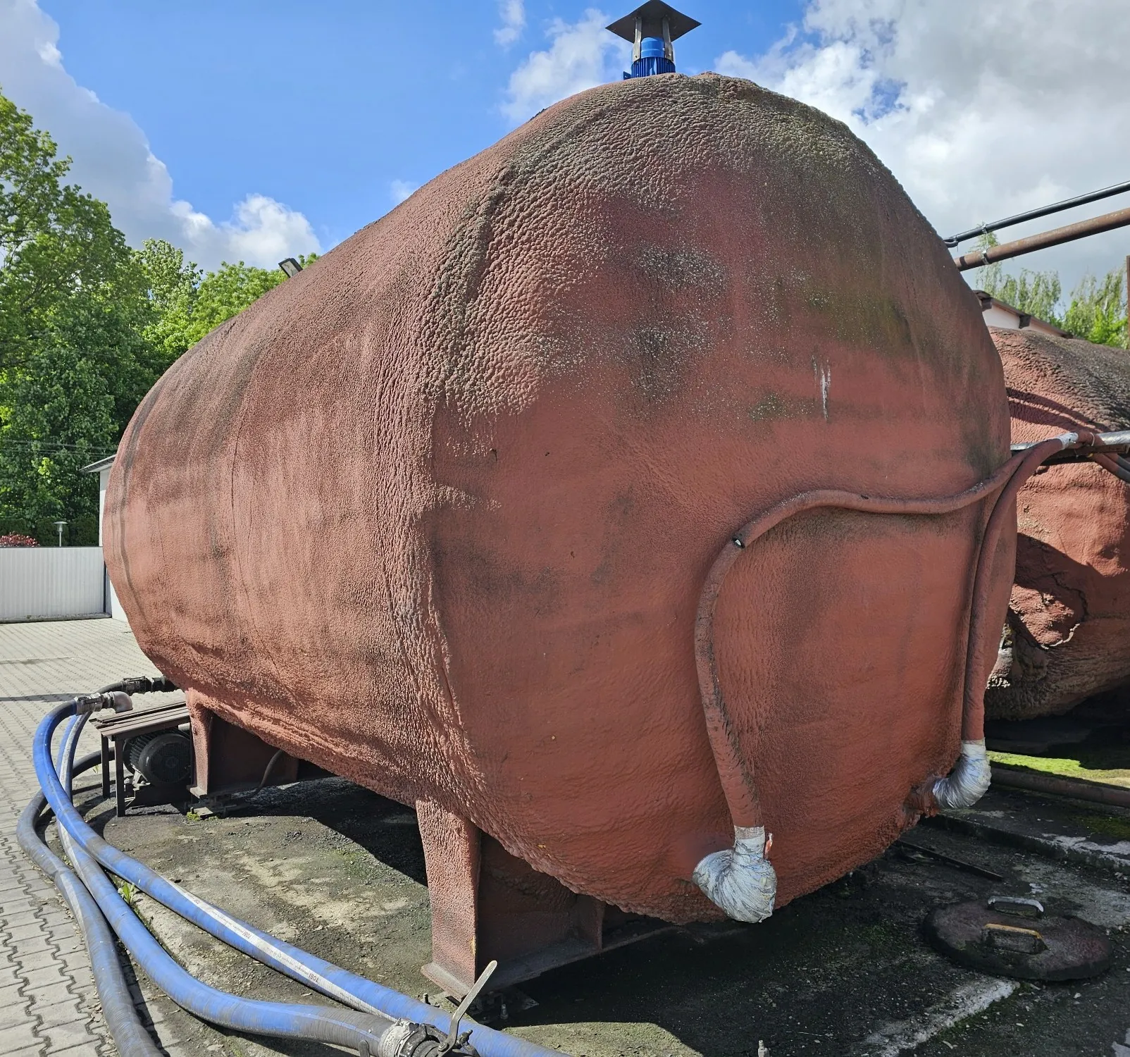 Duży, poziomy zbiornik przemysłowy o zaokrąglonym kształcie, pomalowany na kolor ciemnoczerwony z chropowatą fakturą powierzchni. Zbiornik spoczywa na metalowych podporach. Z jego boku odchodzą rury i przewody, a u dołu widoczne są niebieskie węże przemysłowe. W tle widać zielone drzewa oraz niebo z białymi chmurami.