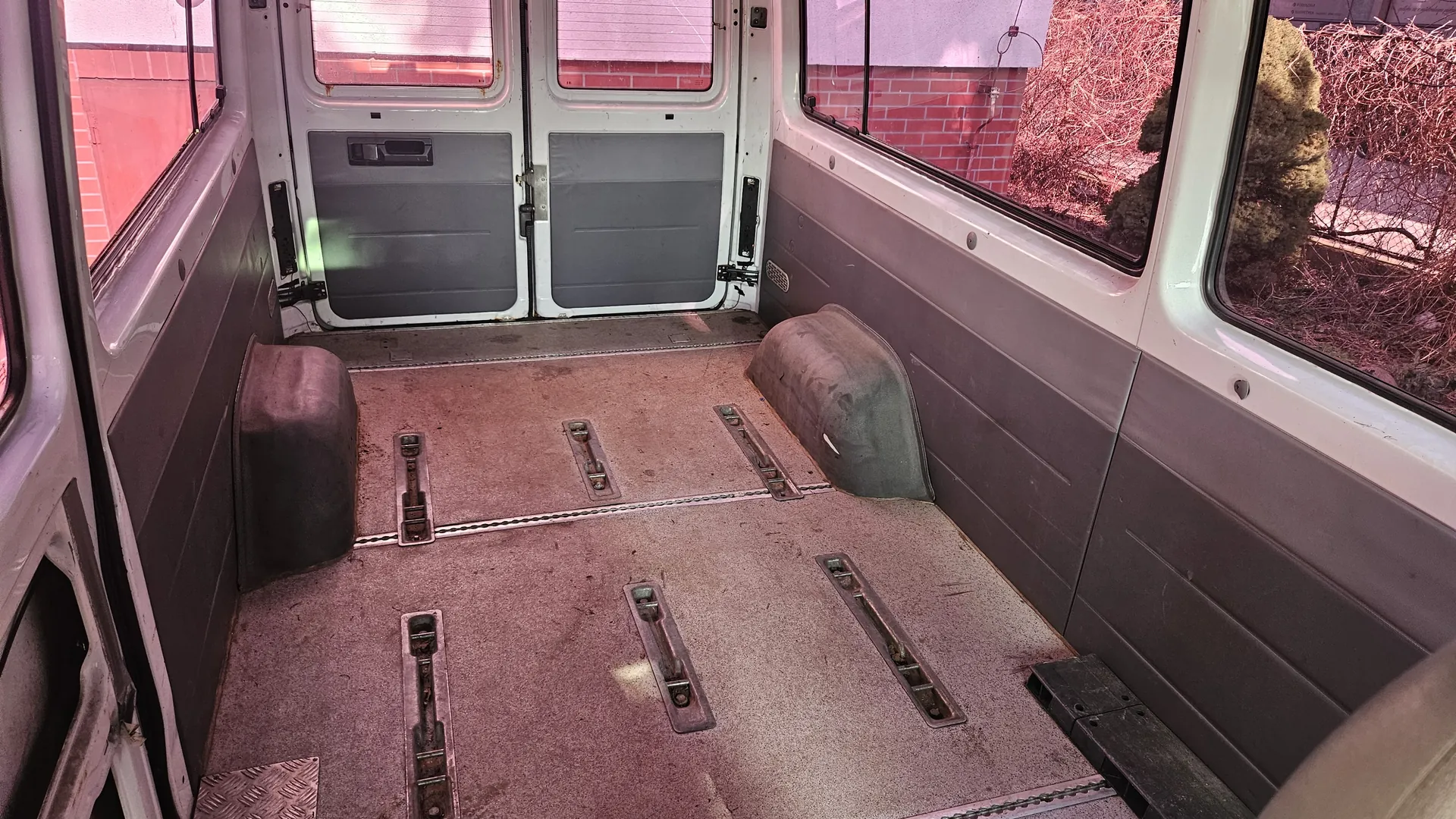 Wnętrze pustego furgonetki z widocznymi metalowymi uchwytami na podłodze. Drzwi tylne są zamknięte, a przez boczne okna widać ceglaną ścianę i suchą roślinność. Wnętrze posiada szare i białe panele, a podłoga jest brudna z metalowymi elementami mocującymi.