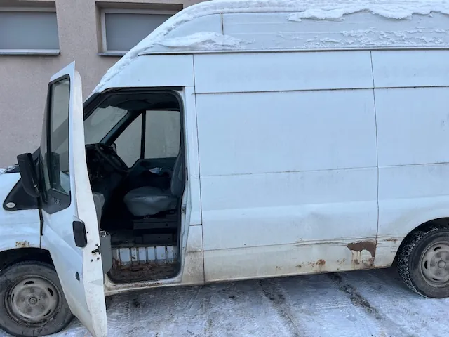Biały furgon zaparkowany na zaśnieżonym podłożu. Przednie lewe drzwi kierowcy są otwarte, odsłaniając wnętrze kabiny z fotelem i kierownicą. Na dachu pojazdu znajduje się warstwa śniegu. Dolna część nadwozia samochodu jest zabrudzona i posiada widoczne ślady rdzy. W tle widać fragment jasnej ściany budynku z dwoma oknami.