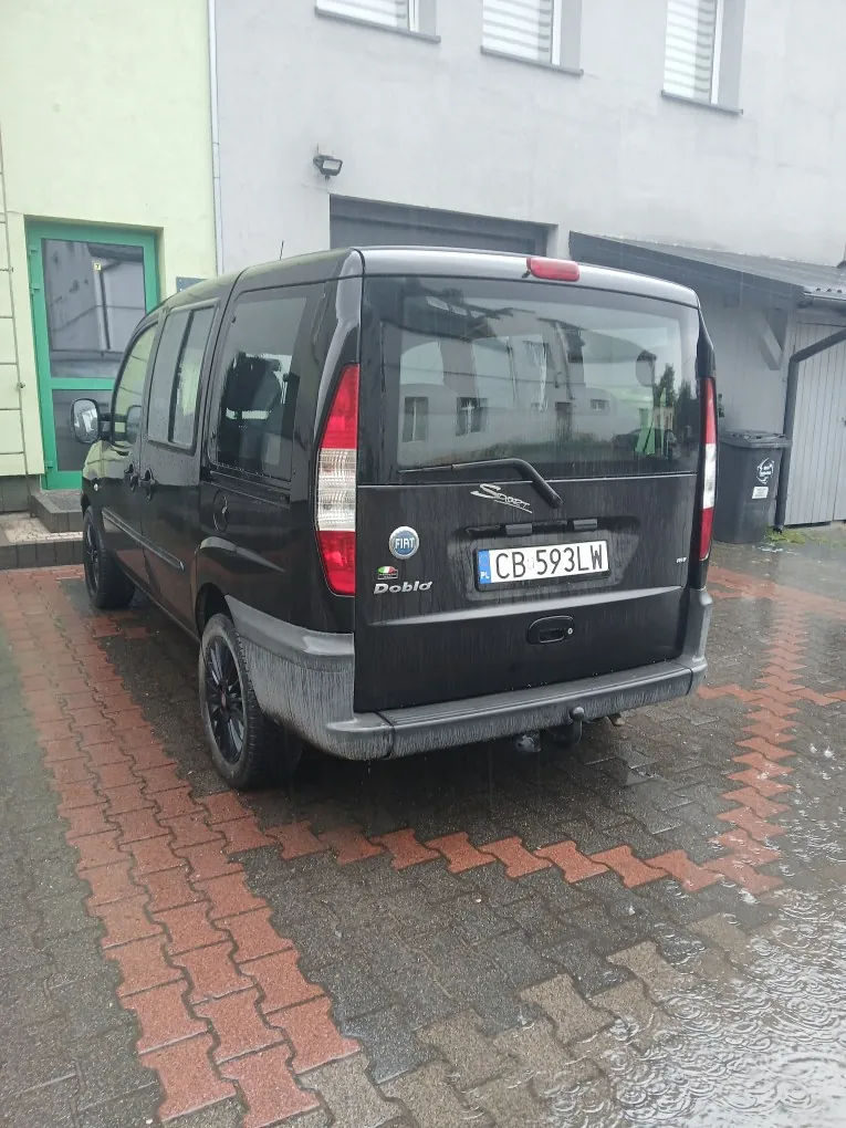 Czarny samochód dostawczy marki Fiat Doblo zaparkowany tyłem na mokrej kostce brukowej przed szarym budynkiem. Na klapie bagażnika widnieją napisy 'FIAT', 'Doblo' oraz tablica rejestracyjna o numerze 'CB 593LW'. W tle widoczny jest budynek z jasną elewacją, zielone drzwi wejściowe oraz czarny kosz na śmieci.