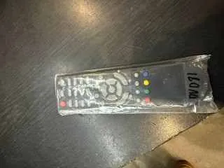 Zdjęcie przedstawia nowy pilot w przezroczystym plastikowym opakowaniu leżący na ciemnej powierzchni. Pilot posiada okrągły pad nawigacyjny, kolorowe przyciski (czerwony, zielony, żółty, niebieski) oraz dodatkowe przyciski. Na opakowaniu widoczny jest pionowy napis '150m'.