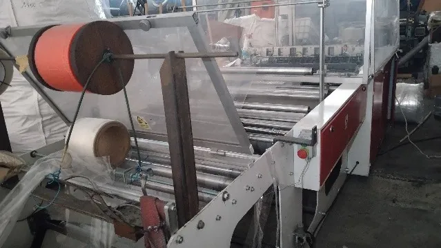 Maszyna przemysłowa do pakowania lub owijania z rolkami i prowadnicami. Po lewej stronie znajduje się duża, pomarańczowa rolka materiału zamontowana na metalowym wałku, poniżej niej mniejsza, biała rolka. Maszyna posiada białe metalowe ramy, przezroczyste osłony z tworzywa oraz system metalowych wałków prowadzących. Po prawej stronie widoczny jest biały panel z czerwonym akcentem i zielonym przyciskiem sterującym.