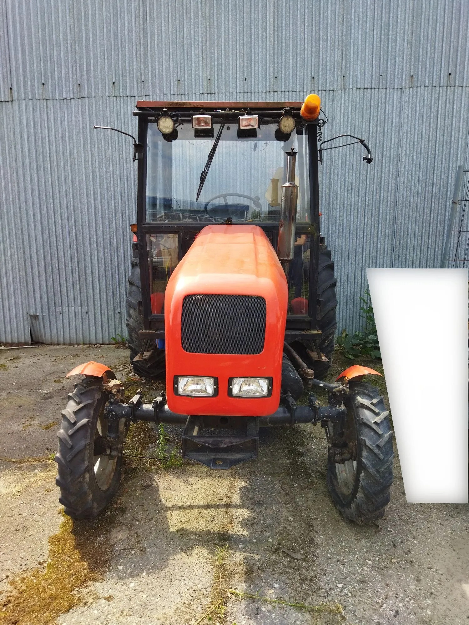 Pomarańczowy traktor z kabiną, zaparkowany na betonowym podłożu przed szarym, falistym blaszanym budynkiem. Maszyna jest zwrócona przodem do widza, posiada duże, ząbkowane opony i dwa reflektory z przodu.