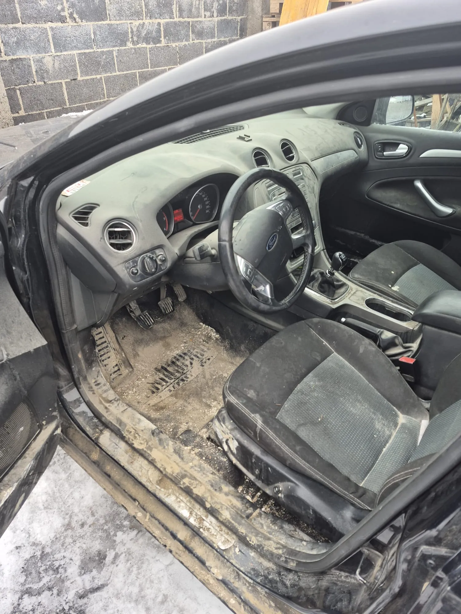 Wnętrze zapuszczonego samochodu z otwartą drzwiami. Brudne siedzenia, zanieczyszczona deska rozdzielcza z kierownicą Ford, podłoga pokryta kurzem i śmieciem. Widoczne pedały, zegary oraz tło z ceglaną ścianą i śniegiem na zewnątrz.