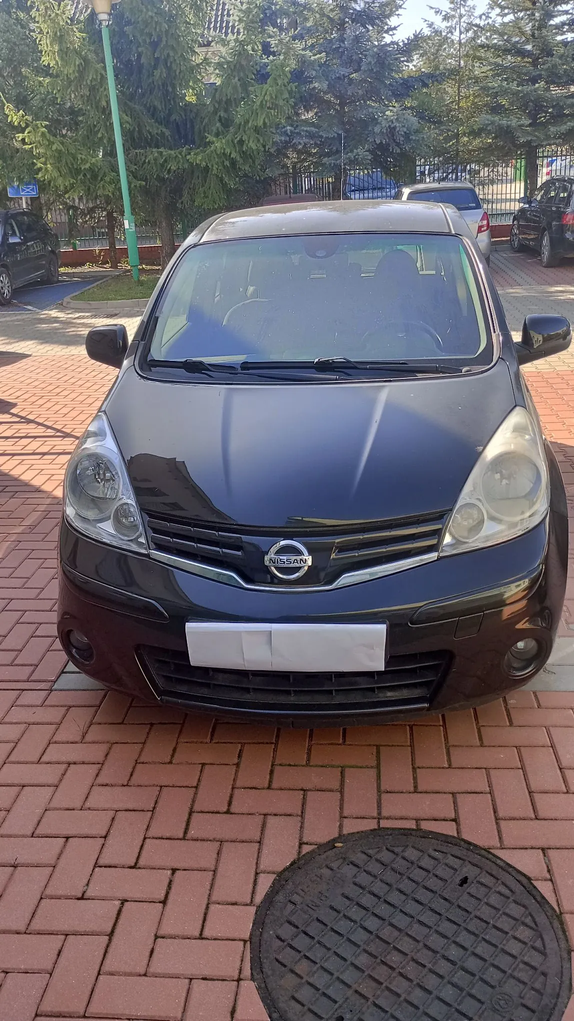 Czarny samochód marki Nissan zaparkowany przodem na czerwonym bruku. W tle widoczne są zielone drzewa iglaste oraz inne zaparkowane pojazdy. Tablica rejestracyjna samochodu jest zakryta białym prostokątem.