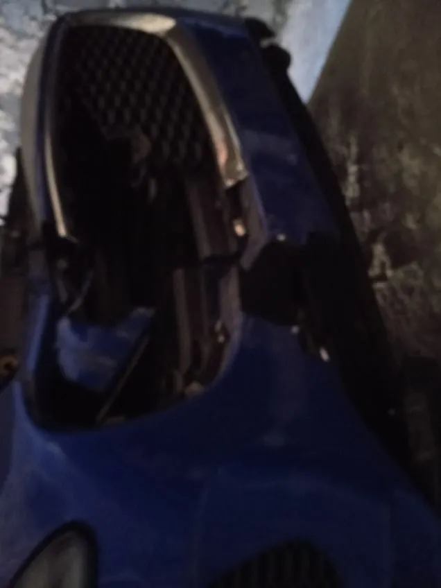 Zbliżenie na przód niebieskiego samochodu z poważnym uszkodzeniem. Brakuje przedniego zderzaka, widoczna jest uszkodzona atrapa chłodnicy i elementy wewnętrzne. Tło stanowi betonowa ściana i podłoga.