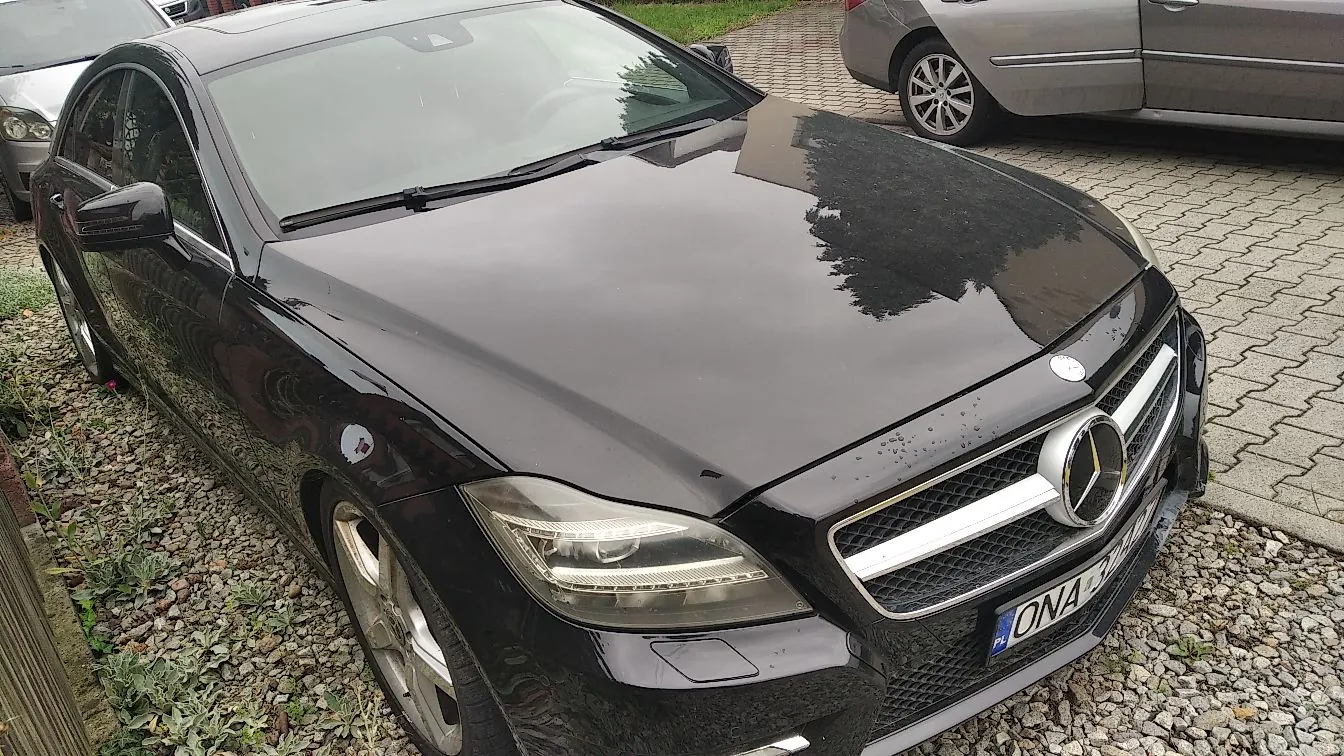 Czarny samochód osobowy marki Mercedes-Benz zaparkowany na podjeździe z kostki brukowej i żwiru. Widoczny jest przód i bok auta, charakterystyczne logo gwiazdy na grillu oraz fragment tablicy rejestracyjnej z końcówką 'ONA' i oznaczeniem 'PL'. W tle widoczny jest fragment innego, srebrnego samochodu.