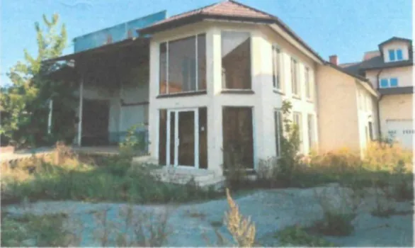 Zdjęcie dwukondygnacyjnego, jasnego budynku o nowoczesnej architekturze z dużymi przeszkleniami na fasadzie. Budynek posiada spadzisty dach kryty ciemną dachówką. Przed domem znajduje się nieuporządkowany teren z niską roślinnością i żwirem. W tle po prawej stronie widoczny jest fragment innego budynku.