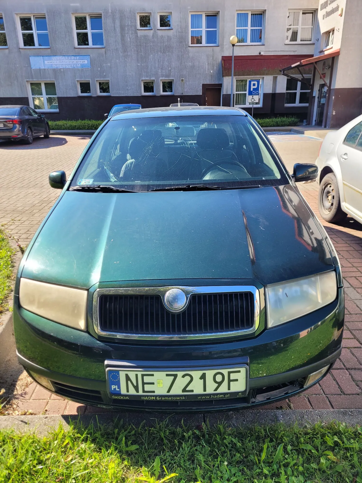 Zielony samochód osobowy marki Skoda zaparkowany na kostce brukowej. Widoczny jest przód pojazdu z tablicą rejestracyjną o numerze NE 7219F. W tle znajduje się szary budynek z oknami oraz fragment trawnika.