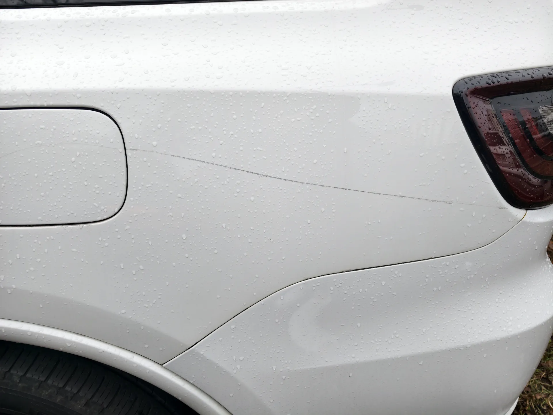 Biały tył samochodu z widocznym zbiornikiem paliwa po lewej stronie, czerwonym światłem tylnym po prawej stronie oraz kroplami wody na karoserii. Na boku pojazdu widoczna jest cienka, pozioma rysa. Część opony i trawy w tle.