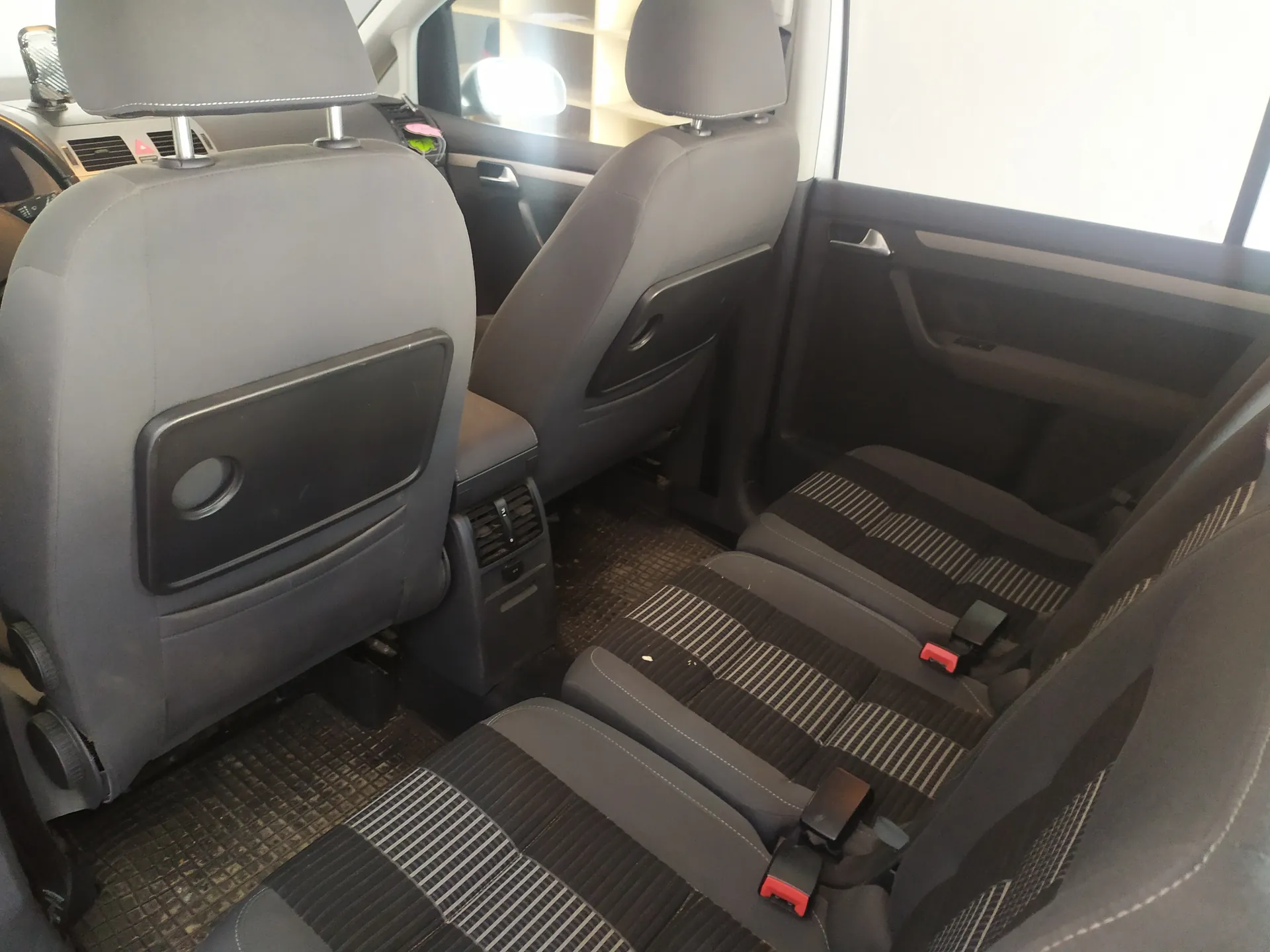 Wnętrze samochodu z widokiem na tylne siedzenia. Widoczne są dwa przednie fotele z podłokietnikami i zagłówkami, a także tylne siedzenia z paskowanym wzorem w kolorze szaro-czarnym. Na siedzeniach znajdują się czerwone klamry pasów bezpieczeństwa. Podłoga pokryta jest matą z siateczkowym wzorem. Po prawej stronie widoczna jest czarna drzwi samochodu z uchwytem i oknem. W tle przez przednią szybę widać fragment budynku z półkami.