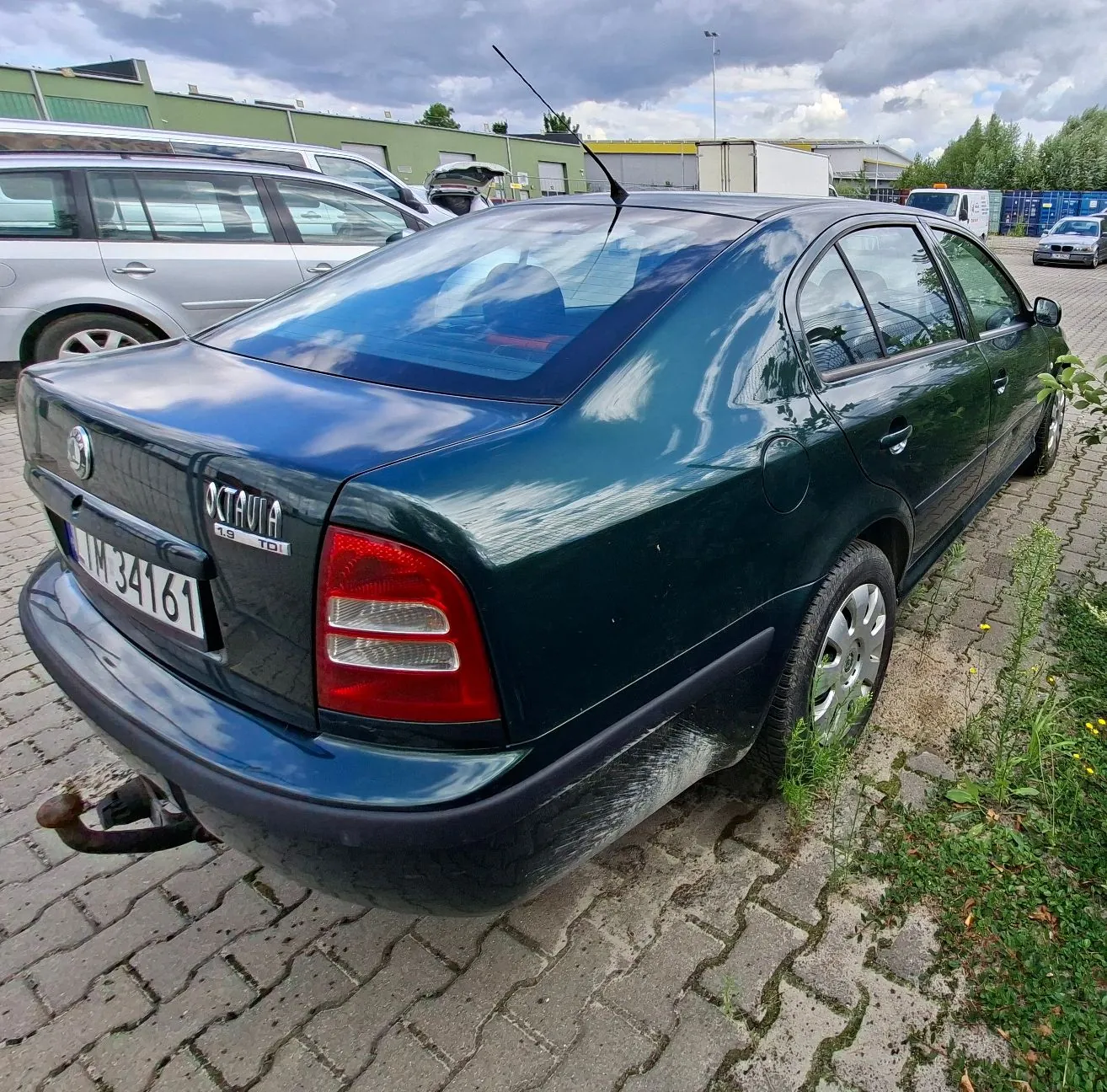 Ciemnozielony samochód osobowy marki Skoda Octavia zaparkowany na kostce brukowej. Widoczny jest tył i prawy bok pojazdu. Na klapie bagażnika znajdują się napisy 'OCTAVIA' oraz '1.9 TDI'. Tablica rejestracyjna ma numer 'IM 34161'. Samochód posiada hak holowniczy. W tle widoczne są inne zaparkowane samochody oraz budynki przemysłowe pod zachmurzonym niebem.