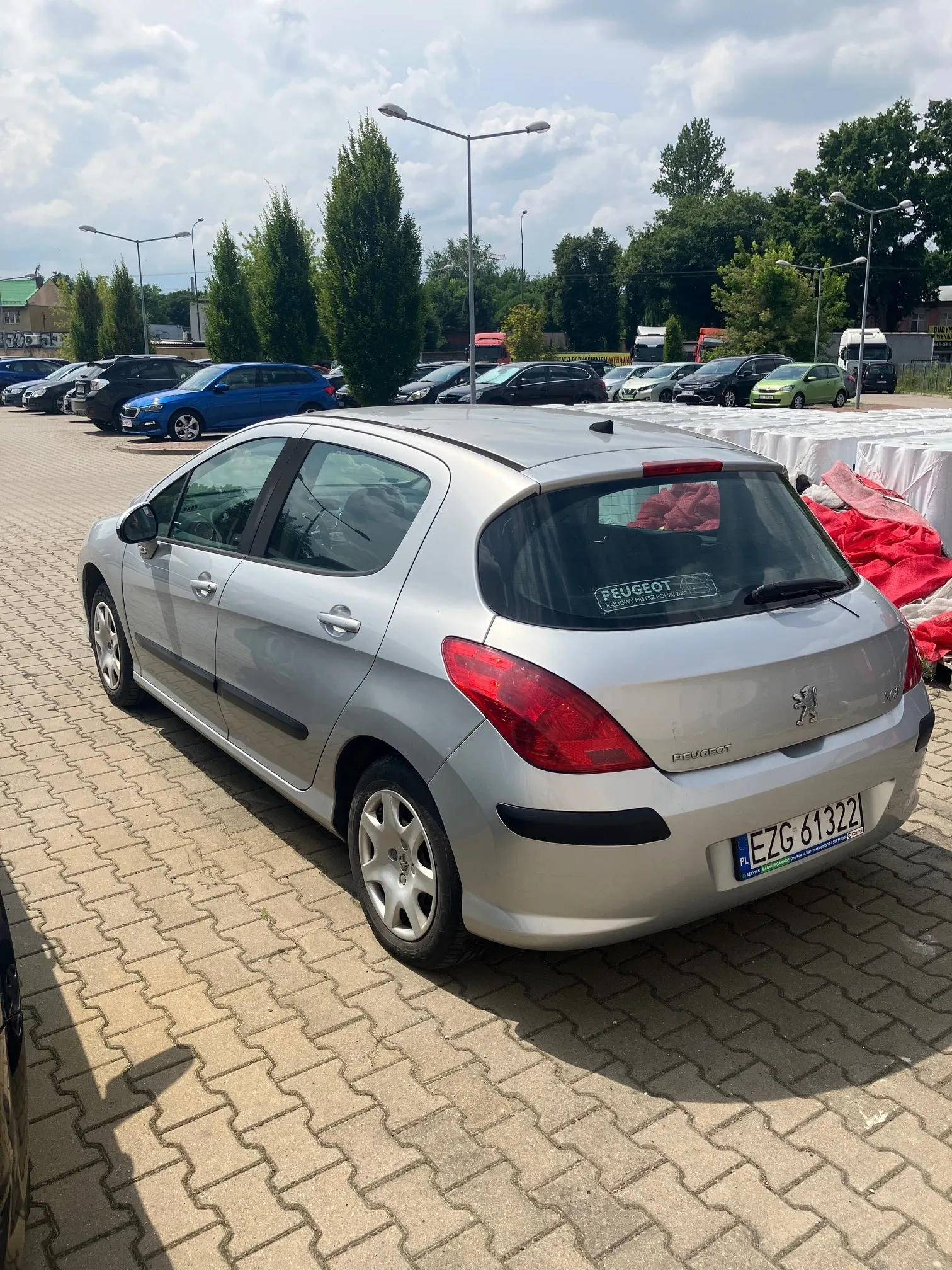 Srebrny samochód marki Peugeot w nadwoziu hatchback zaparkowany na placu z kostki brukowej. Pojazd jest widoczny z tyłu i z boku. Na tylnej szybie znajduje się naklejka z napisem 'PEUGEOT'. Tablica rejestracyjna samochodu to EZG 61322. W tle widoczne są inne zaparkowane samochody w różnych kolorach, zielone drzewa oraz latarnie uliczne pod jasnym, lekko zachmurzonym niebem.