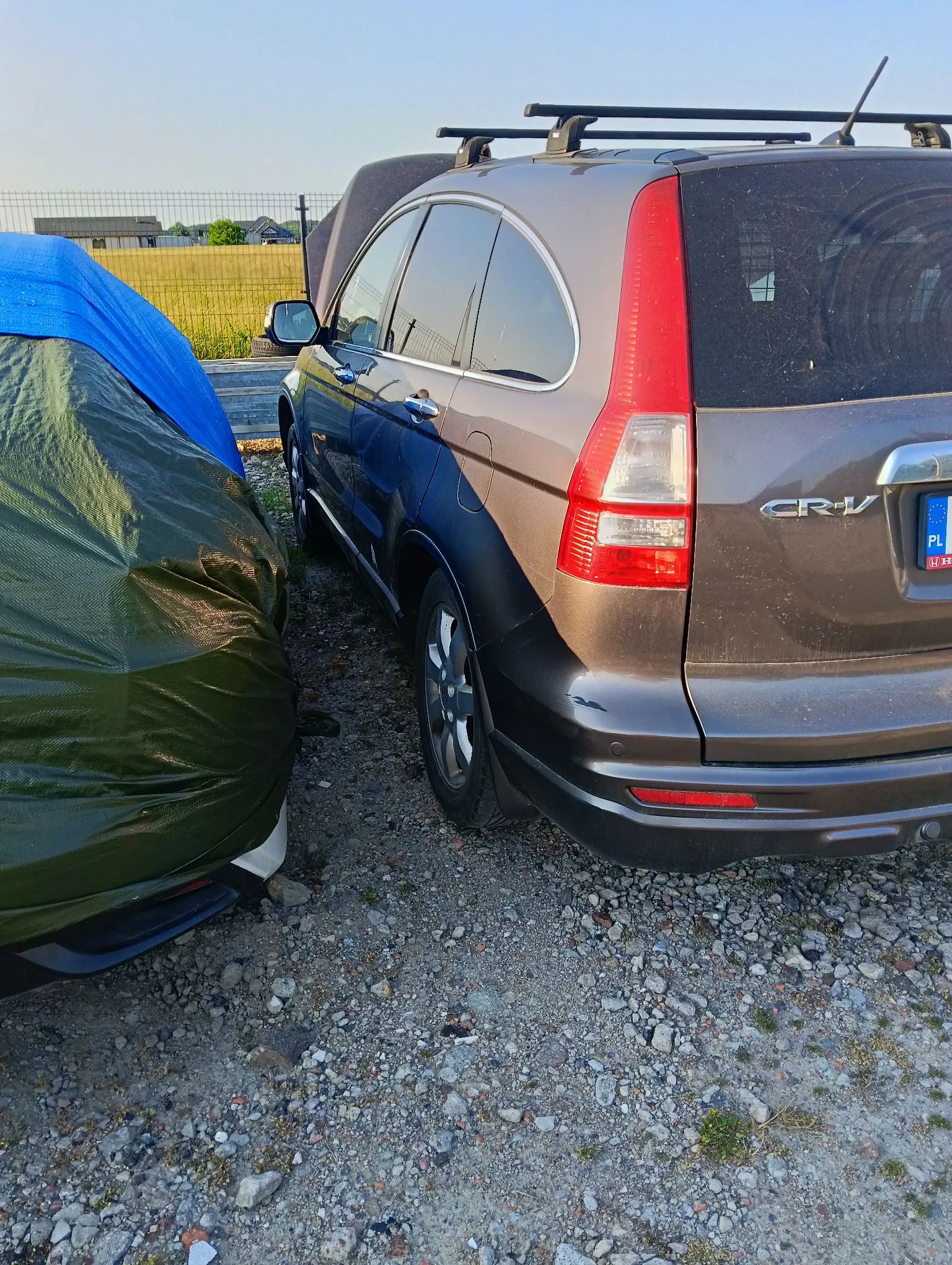 Zdjęcie przedstawiające tył i bok brązowego samochodu marki Honda CR-V zaparkowanego na żwirowym podłożu. Na dachu samochodu zamontowany jest bagażnik. Obok, po lewej stronie, znajduje się inny pojazd przykryty ciemnozieloną i niebieską plandeką. W tle widoczne jest pole i niskie zabudowania pod jasnym niebem.