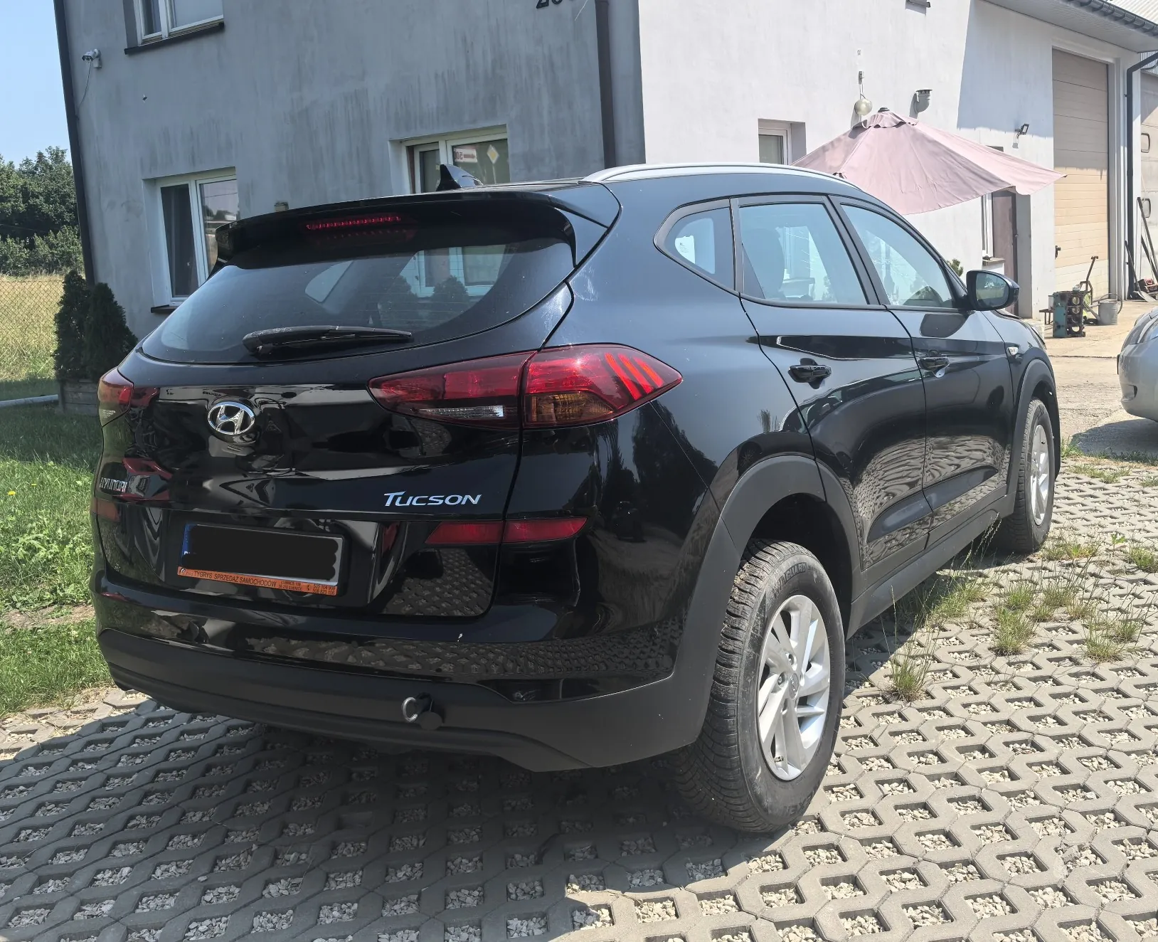 Czarny samochód marki Hyundai Tucson zaparkowany tyłem na kostce brukowej przed białym budynkiem. Widoczny jest tylny zderzak, lampy, logo Hyundai oraz napis TUCSON. W tle znajduje się dom z oknami oraz rozłożony różowy parasol ogrodowy.