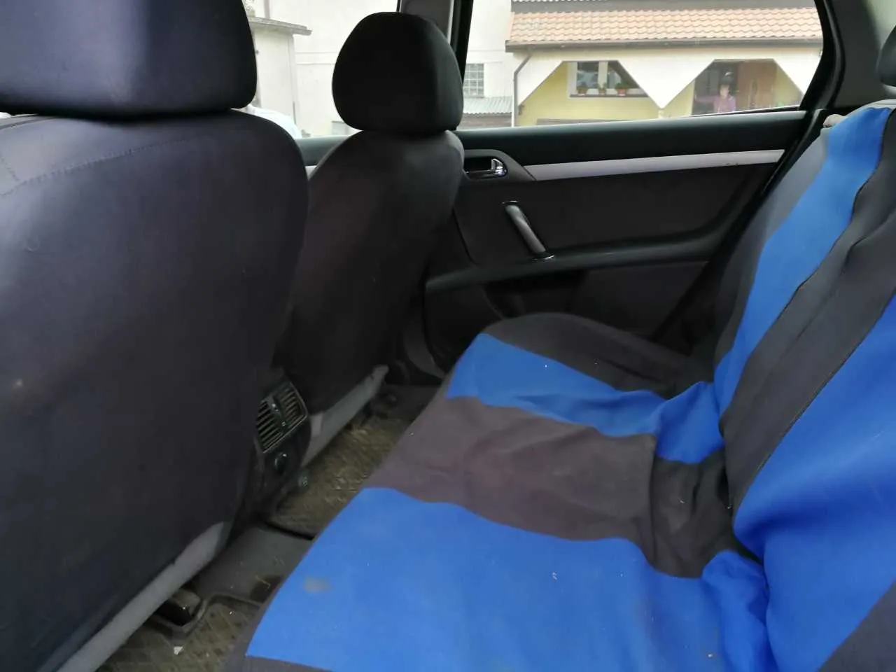 Wnętrze samochodu widziane z tyłu. Widoczne są dwa przednie siedzenia: lewe z ciemnym pokrowcem, prawe z niebiesko-czarnym pasowanym pokrowcem. Drzwi po stronie pasażera są otwarte, przez okno widać budynek z żółtą elewacją, czerwoną dachówką i osobą stojącą w drzwiach. Wnętrze samochodu ma ciemne panele, a podłoga jest wyłożona matą. Na siedzeniu z niebieskim pokrowcem widoczne są ślady zabrudzeń.