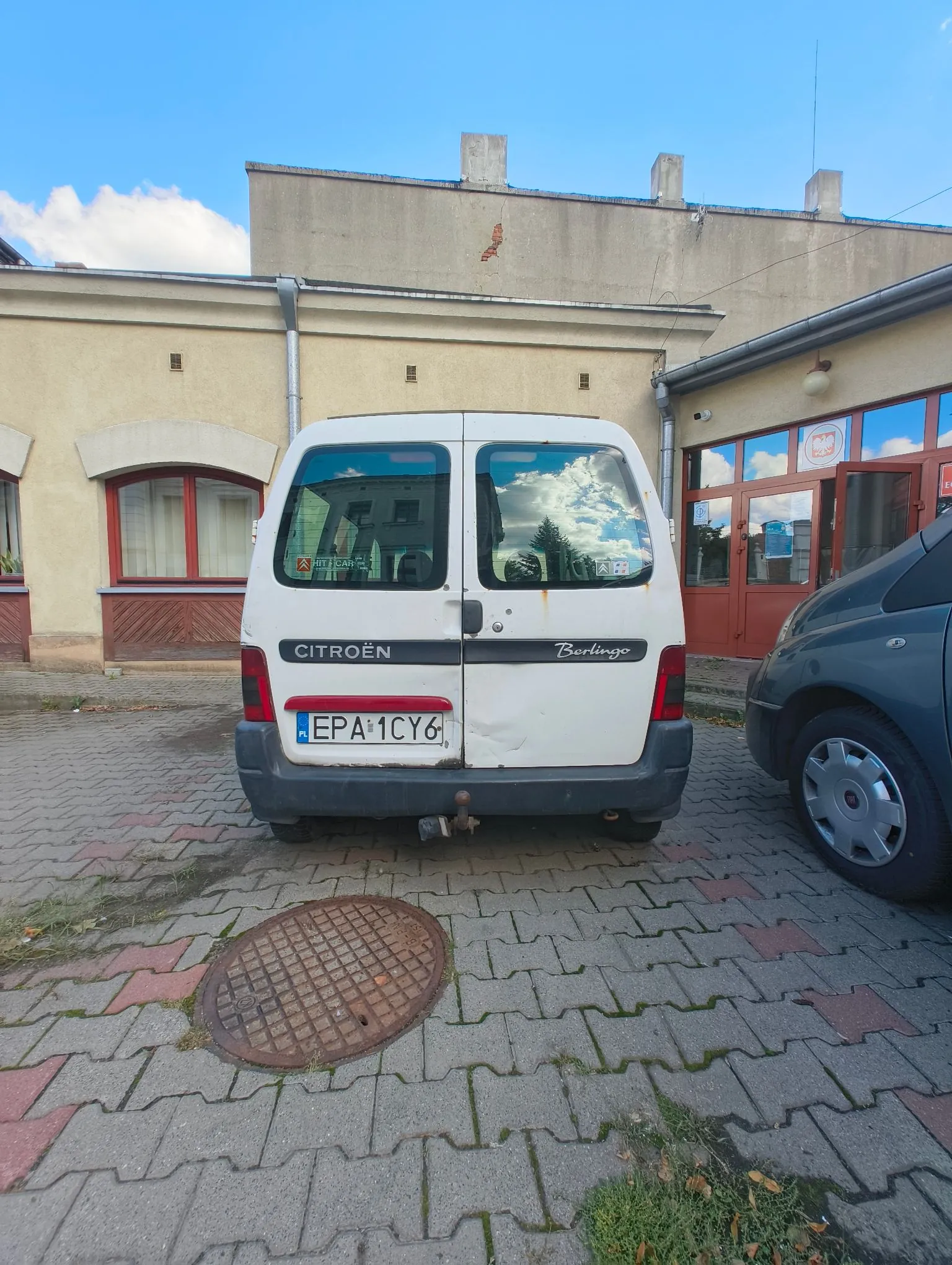 Widok z tyłu białego samochodu dostawczego marki Citroën Berlingo zaparkowanego na kostce brukowej przed niskim, jasnym budynkiem. Pojazd ma tablicę rejestracyjną o numerze EPA 1CY6 z oznaczeniem kraju AL. Obok stoi fragment ciemnego samochodu. W tle widoczny jest budynek z oknami i czerwonymi drzwiami, nad którymi znajduje się godło Polski.