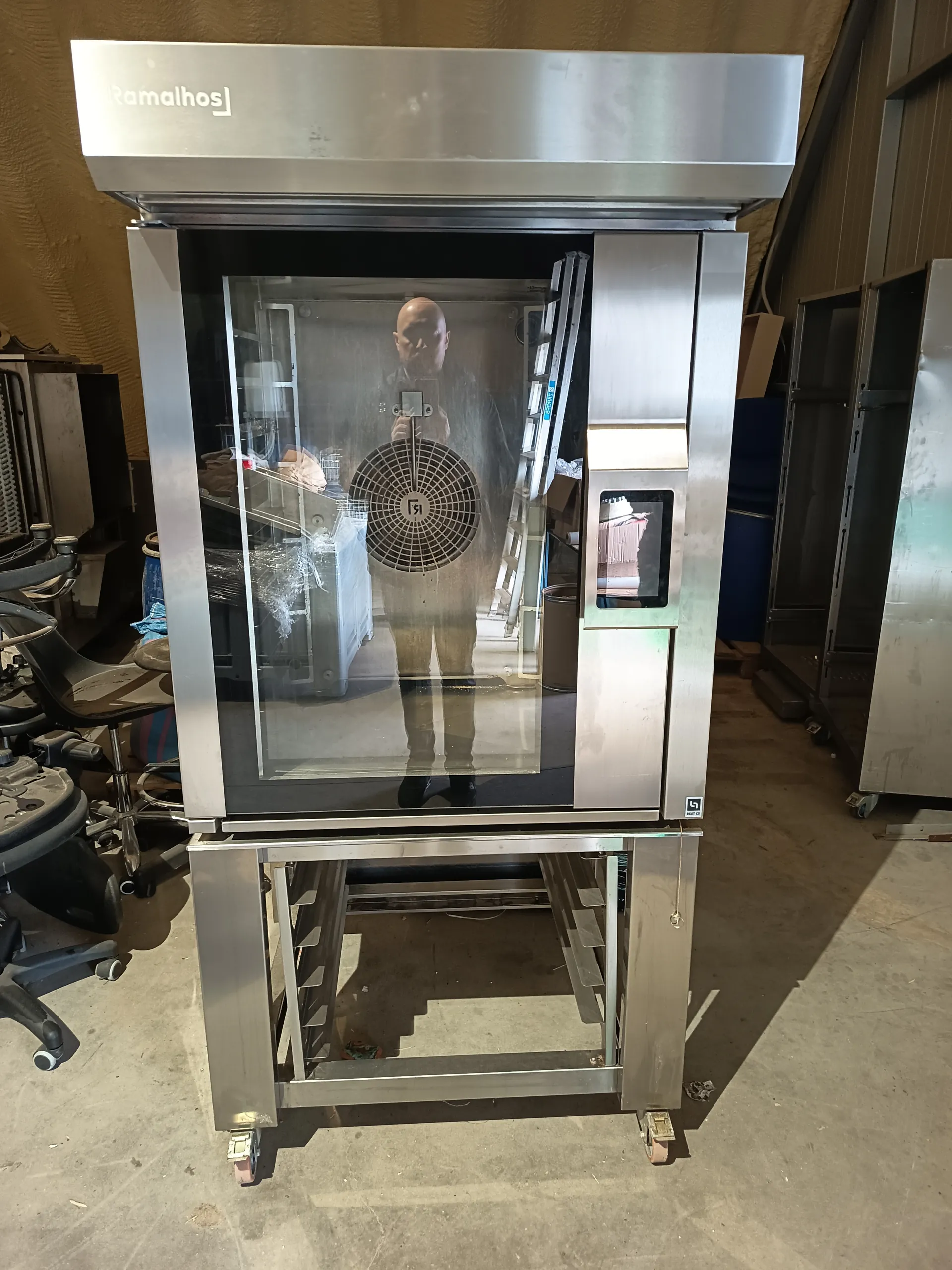 Stalowa maszyna przemysłowa marki Ramalhos z przezroczystą drzwiami i okrągłym wentylatorem oznaczonym numerem 51 w środku. Na drzwiach widoczne odbicie osoby fotografującej. Maszyna stoi na kółkach w przemyślnym pomieszczeniu z betonową podłogą. W tle znajdują się czarne krzesła biurowe, drabina, opakowania i inne urządzenia przemysłowe.