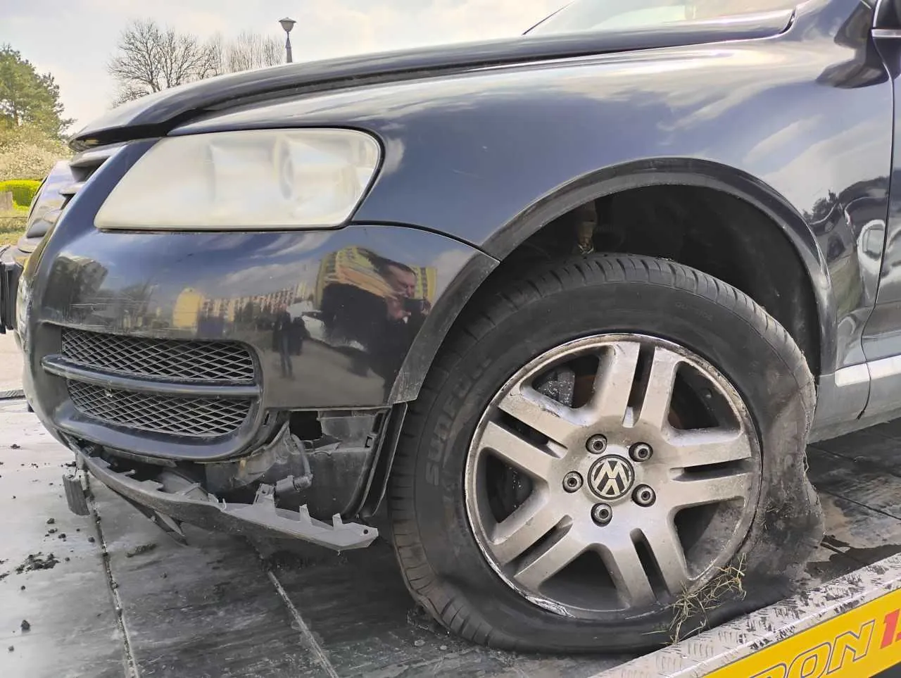 Czarny samochód osobowy marki Volkswagen z widocznymi uszkodzeniami przedniej części. Zniszczony zderzak, pęknięta maska i uszkodzone koło przednie lewe. Na felgach widoczny logo VW. W tle drzewa, latarnia uliczna oraz odbicie budynków i osób na błyszczącej karoserii. Samochód stoi na powierzchni z metalowymi płytkami, częściowo widoczny żółty znak z napisem 'ON 1'. Niebo jest częściowo zachmurzone.