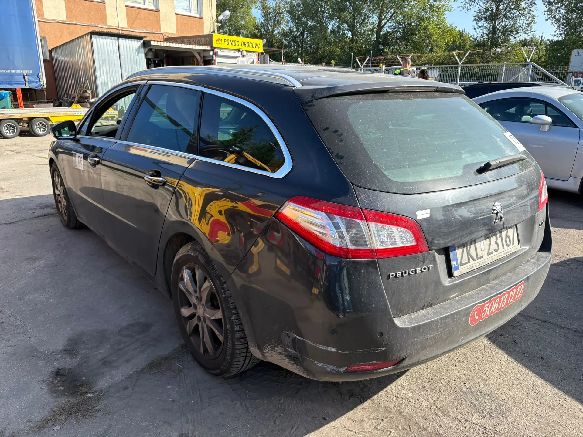 Czarny samochód marki Peugeot w nadwoziu kombi, zaparkowany na piaszczystym placu. Widoczny jest tył i lewy bok pojazdu. Samochód jest zakurzony i posiada uszkodzenia w postaci zarysowań na tylnym lewym zderzaku. Tablica rejestracyjna ma numer ZKL 23167. Na klapie bagażnika widnieje napis PEUGEOT oraz naklejka z numerem telefonu 506 13 12 12. W tle widać budynek z żółtym szyldem z napisem POMOC DROGOWA oraz inny, srebrny samochód.