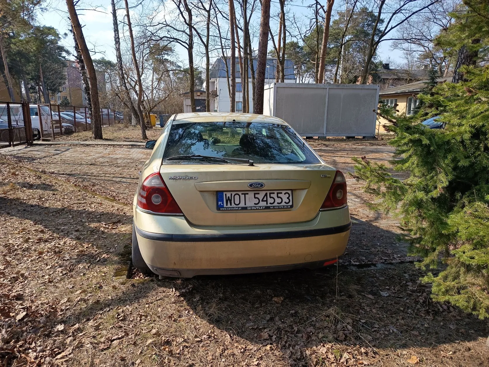 Złoty samochód Ford Mondeo z tablicą rejestracyjną PL WOT 54553 zaparkowany na terenie z suchymi liśćmi. W tle widoczne są drzewa bez liści, budynki, ogrodzenie oraz inne zaparkowane pojazdy. Na tablicy rejestracyjnej widoczny napis www.caroutlet.pl oraz numer telefonu. Pogoda słoneczna, niebo częściowo zachmurzone.