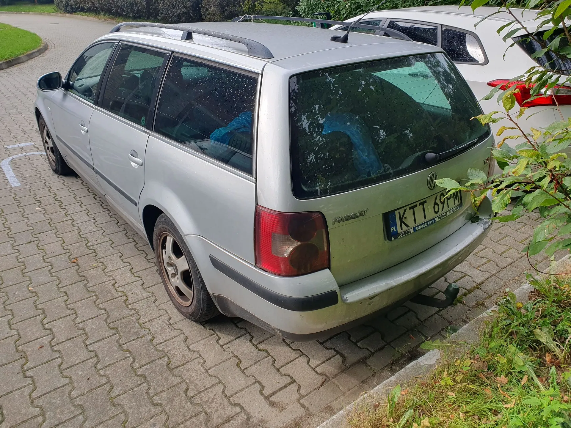 Srebrny samochód marki Volkswagen Passat w wersji kombi, zaparkowany na kostce brukowej. Widoczny jest tył i lewy bok pojazdu. Samochód ma polską tablicę rejestracyjną o numerze KTT 69FM. W tle widać fragment białego samochodu oraz zieloną roślinność.