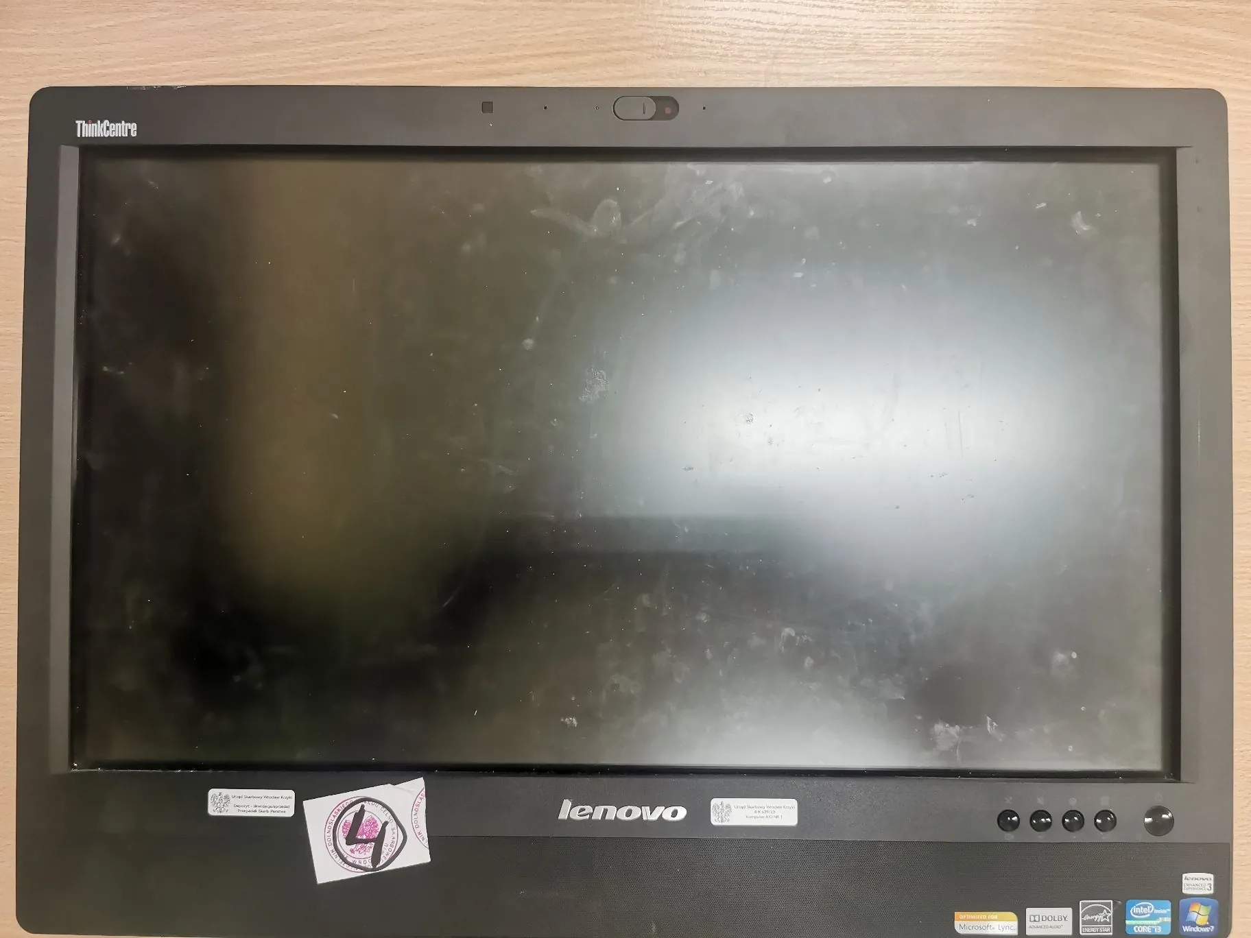 Zdjęcie z przodu czarnego monitora marki Lenovo z serii ThinkCentre. Ekran jest wyłączony i widoczne są na nim liczne smugi oraz zabrudzenia. Na dolnej ramce znajduje się napis 'lenovo' oraz kilka naklejek informacyjnych i certyfikatów, w tym logo Intel Core i5 oraz Windows 7. W lewym górnym rogu ramki widnieje napis 'ThinkCentre'.