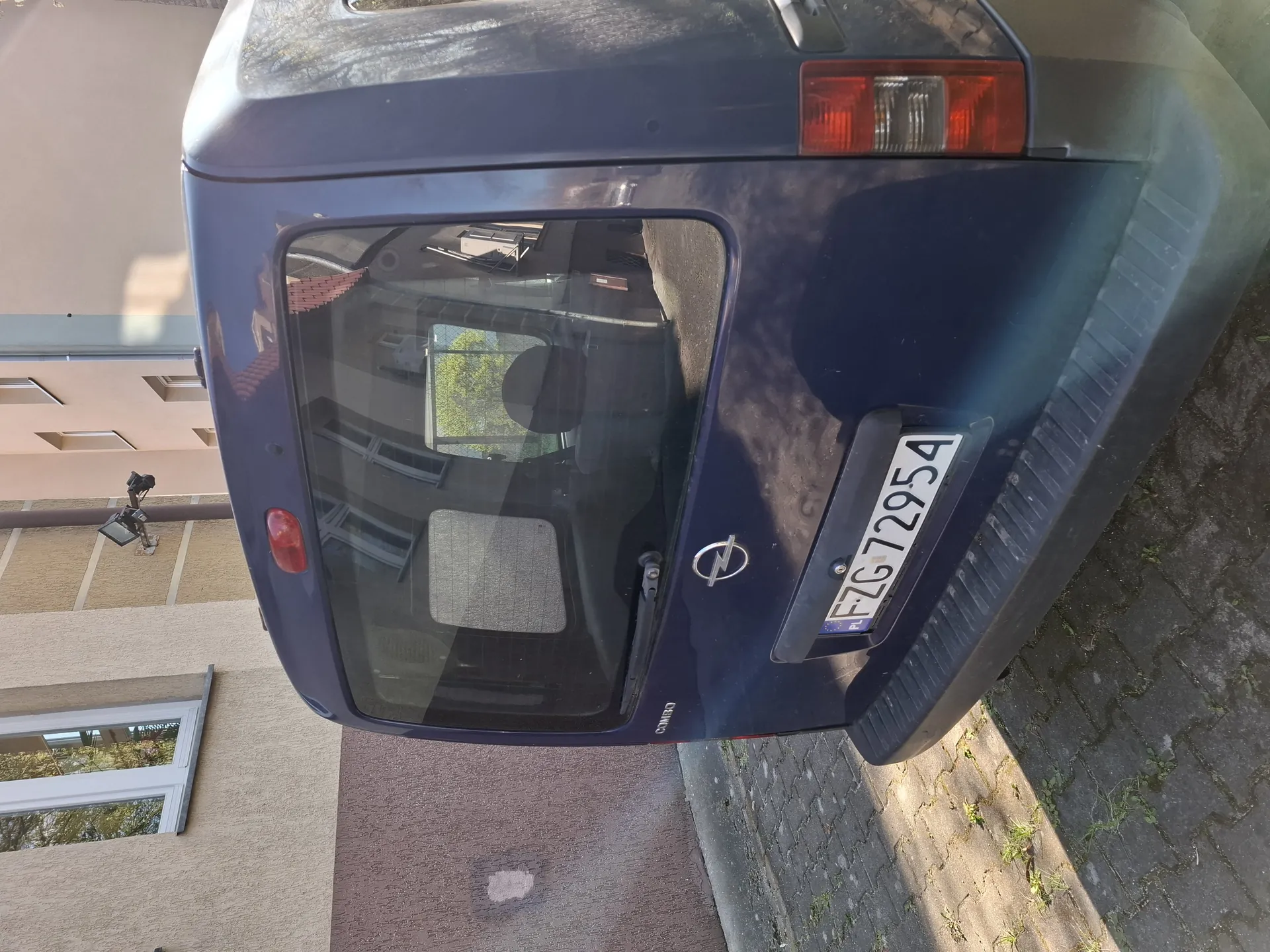 Tylne widok ciemnoniebieskiego samochodu z tablicą rejestracyjną FZG72954, logo marki na bagażniku. Samochód zaparkowany przy budynku o beżowych ścianach z oknami. Widoczne są ceglane chodniki oraz reflektory tylne. W tle drzewa i inne budynki odbijające się w szybie tylnej.