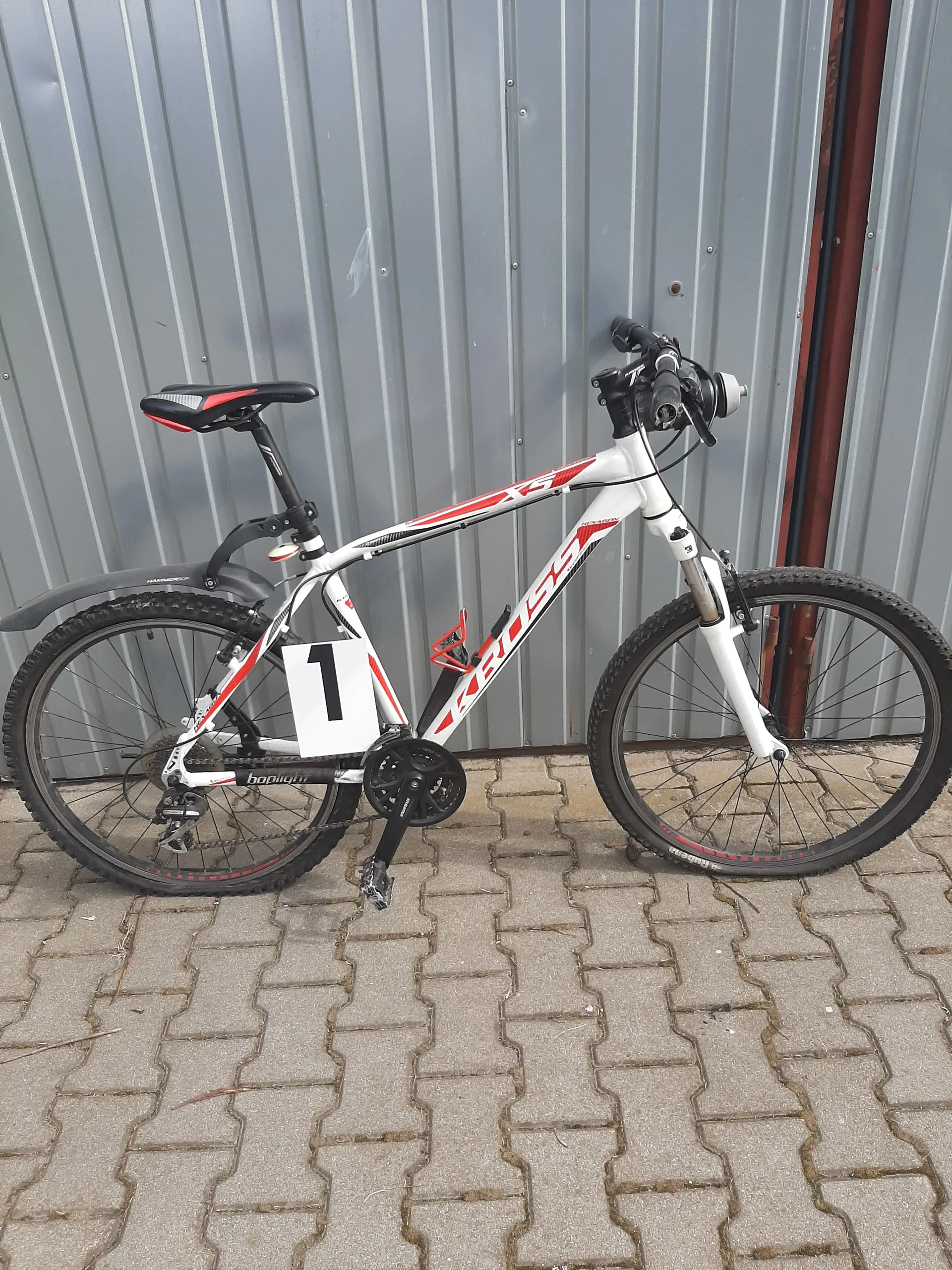 Biały rower z czerwonymi akcentami i napisem 'KROSS' na ramie, z białą tabliczką z czarną cyfrą '1' przymocowaną do ramy. Rower opiera się o szarą metalową ścianę z pionowymi listwami i brukowaną powierzchnię. Widoczne są czarne koła z czerwonymi akcentami, czarno-czerwone siedzisko oraz kierownica z dzwonkiem.