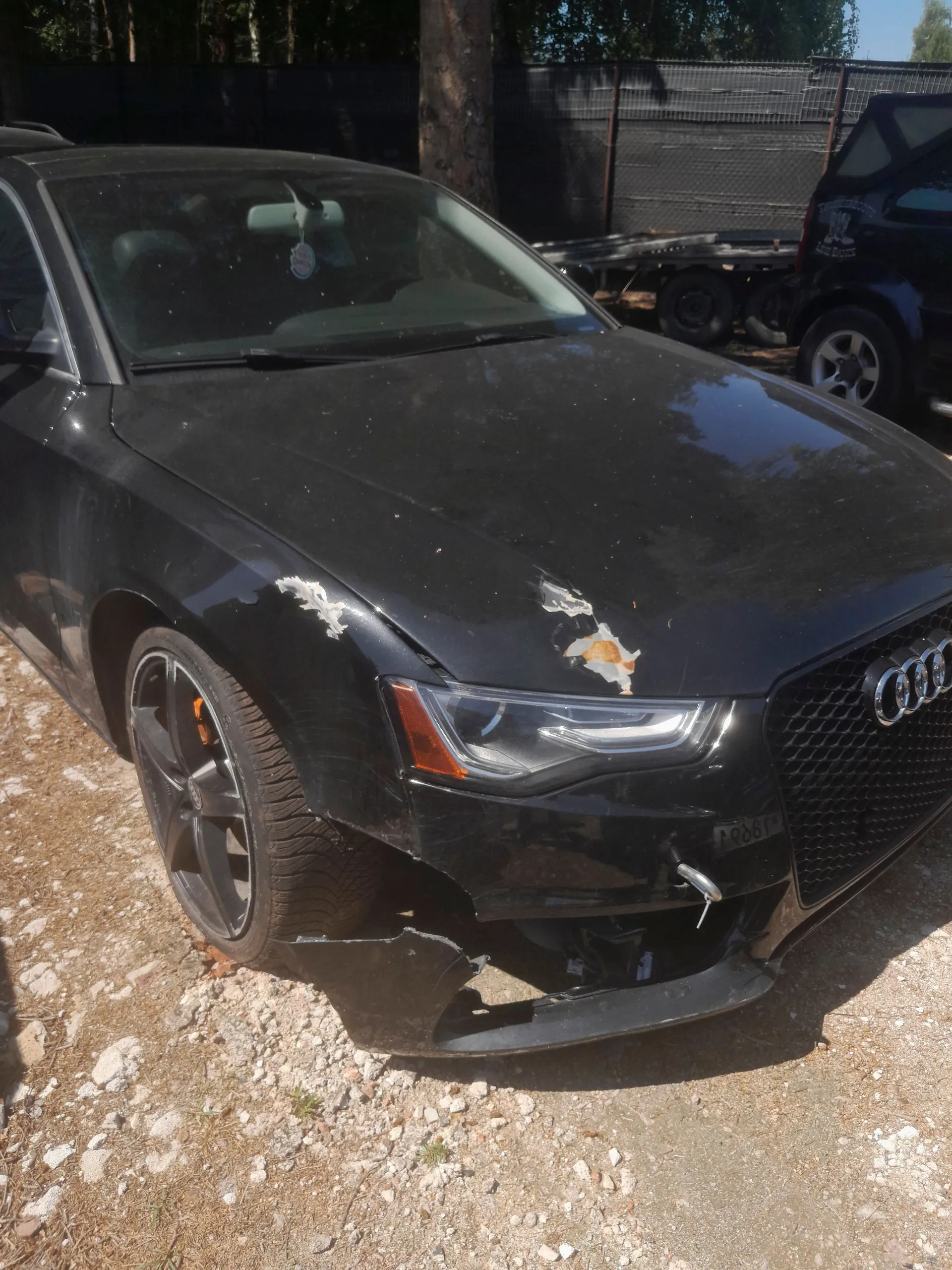 Czarny samochód marki Audi z widocznymi uszkodzeniami przodu. Na masce widoczne są odpryski lakieru i zarysowania, a przedni zderzak jest częściowo oderwany i pęknięty po prawej stronie (patrząc od przodu). Auto stoi na piaszczystym podłożu.