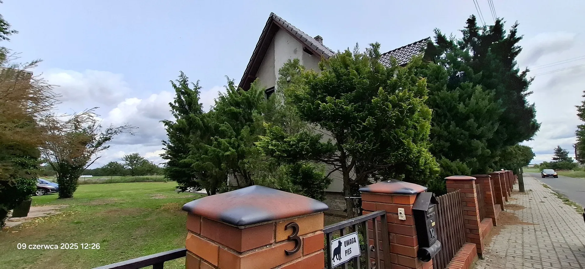 Zdjęcie przedstawia fragment posesji z domem jednorodzinnym w tle, częściowo zasłoniętym przez gęste, zielone krzewy i drzewa iglaste. Na pierwszym planie widoczny jest ceglany słup ogrodzenia z numerem domu '3' oraz tabliczką z napisem 'UWAGA PIES' i sylwetką psa. Obok słupa znajduje się czarna skrzynka pocztowa zamontowana na drewnianym płocie. Po prawej stronie widać chodnik i fragment drogi z jadącym samochodem. W lewym dolnym rogu znajduje się data i godzina: '09 czerwca 2025 12:26'.