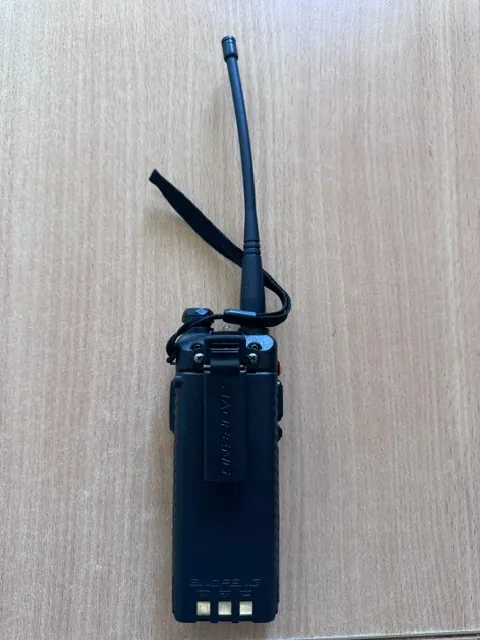 Czarna radiostacja z długą anteną i czarnym paskiem, leżąca na jasnobrązowym, drewnianym blacie. Na obudowie urządzenia widnieje pionowy napis 'BAOFENG' oraz mniejszy napis na dole.