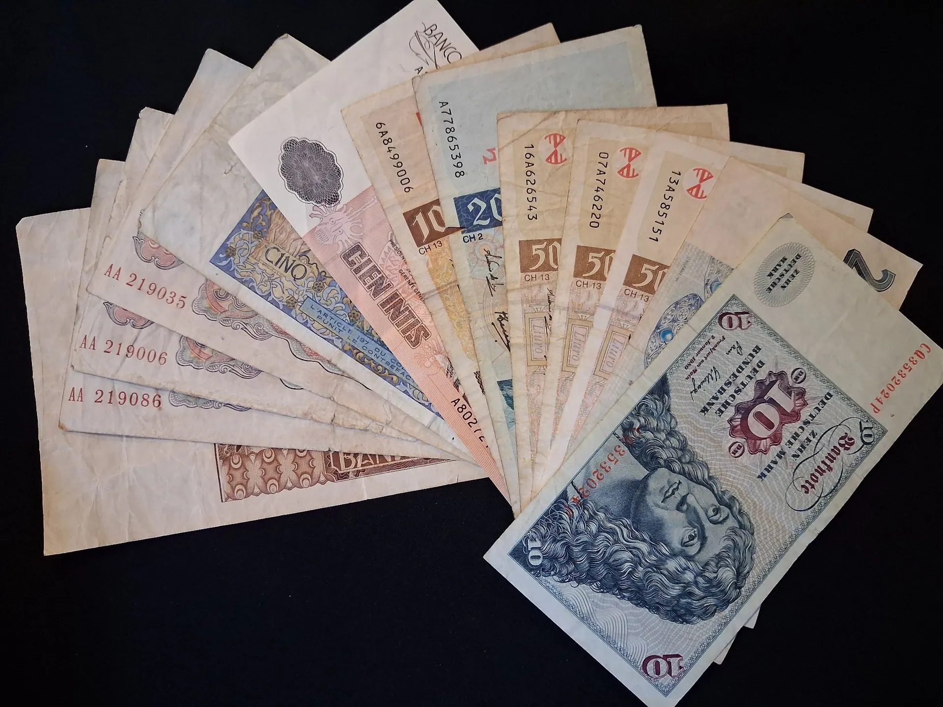 Rozłożony wachlarz starych banknotów z różnych krajów i okresów na czarnym tle. Widoczne są nominały takie jak 10, 20, 50 oraz 100. Na niektórych banknotach widnieją napisy: 'CIEN INTIS', 'DEUTSCHE BANK', 'ZEHN'. Banknoty mają różne kolory: od białych, przez beżowe i niebieskie, aż po różowe. Niektóre z nich są zniszczone i pożółkłe.