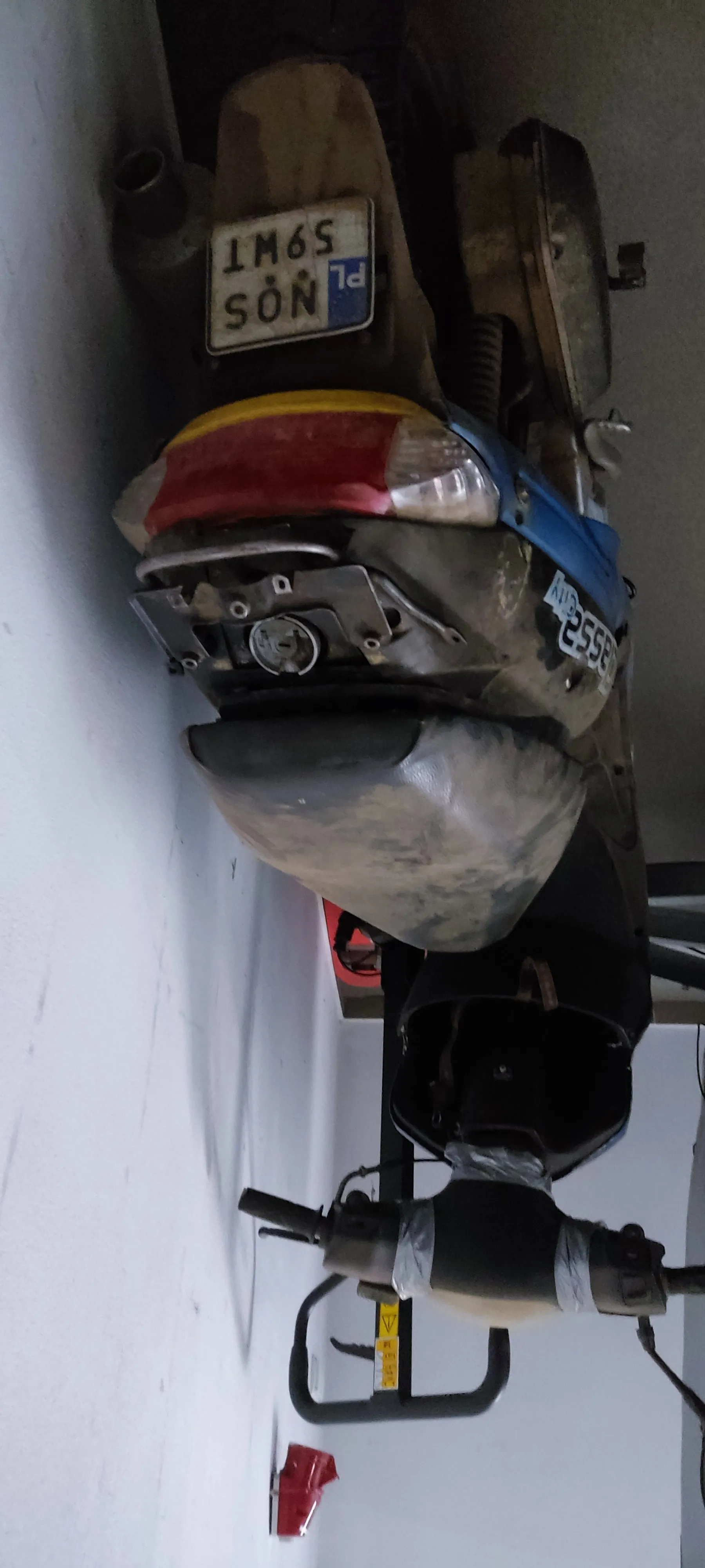 Zdjęcie przedstawia brudny, stary skuter w nieprawidłowej orientacji. Skuter ma czarną siedzisko, niebieską część z napisem "CITY", czerwono-żółty tylny reflektor oraz tablicę rejestracyjną z widocznym tekstem "PL 1M65 S9NT". Na kierownicy znajduje się żółty znak ostrzegawczy, a na siedzisku czarna torba. W tle biała ściana z czerwonym przedmiotem (prawdopodobnie gaśnicą) zamontowanym na ścianie.